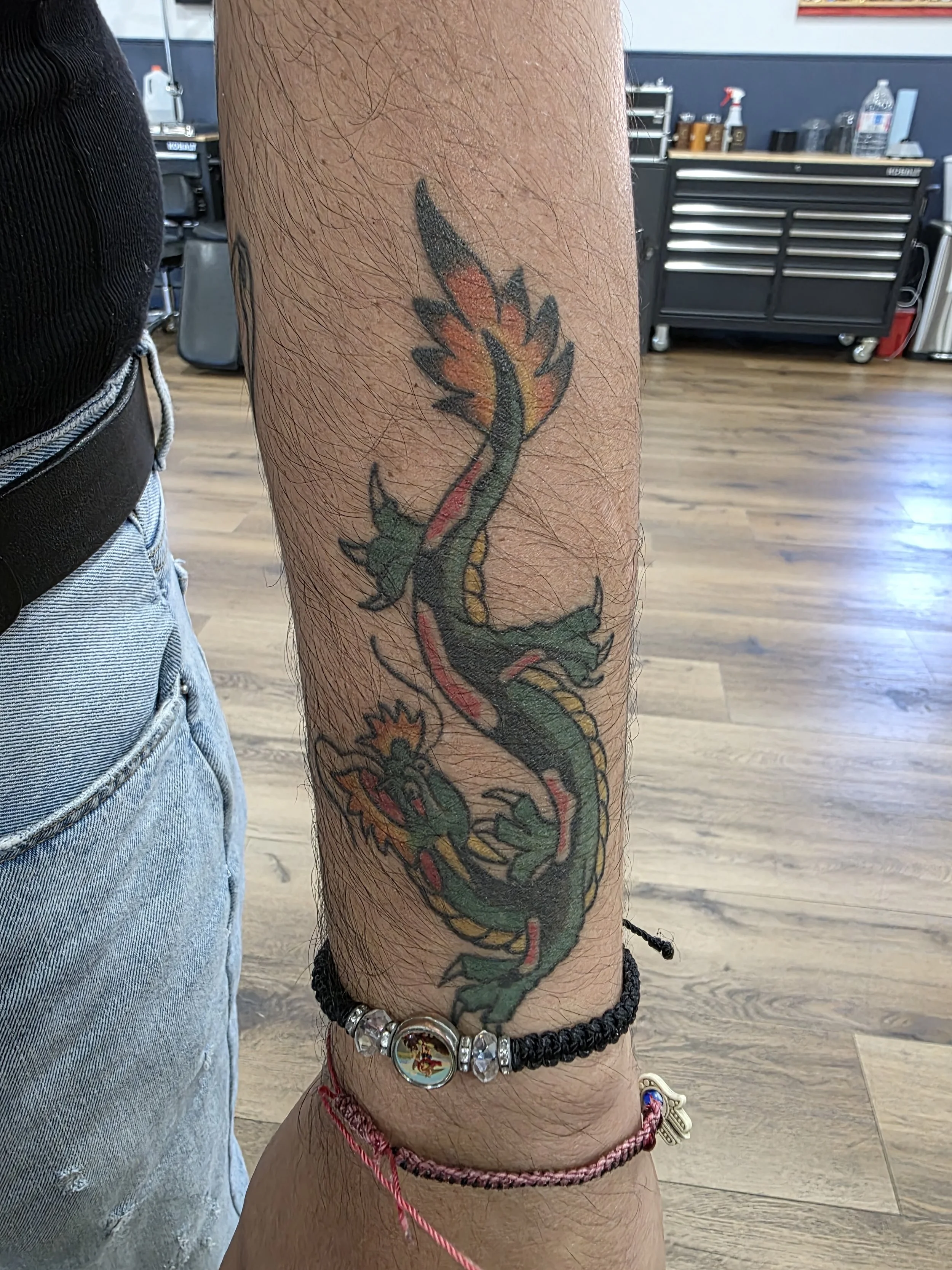 Tattoo of a green dragon with orange flames on the lower leg of a person wearing jeans and bracelets, including a bead bracelet with a round charm, standing in a room with wooden flooring and tools in the background.