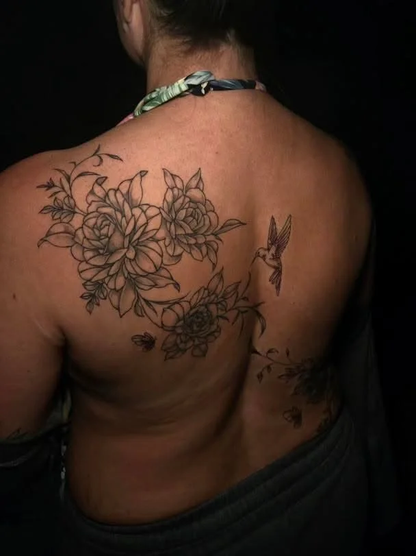 A person's back with a tattoo of flowers and a hummingbird.