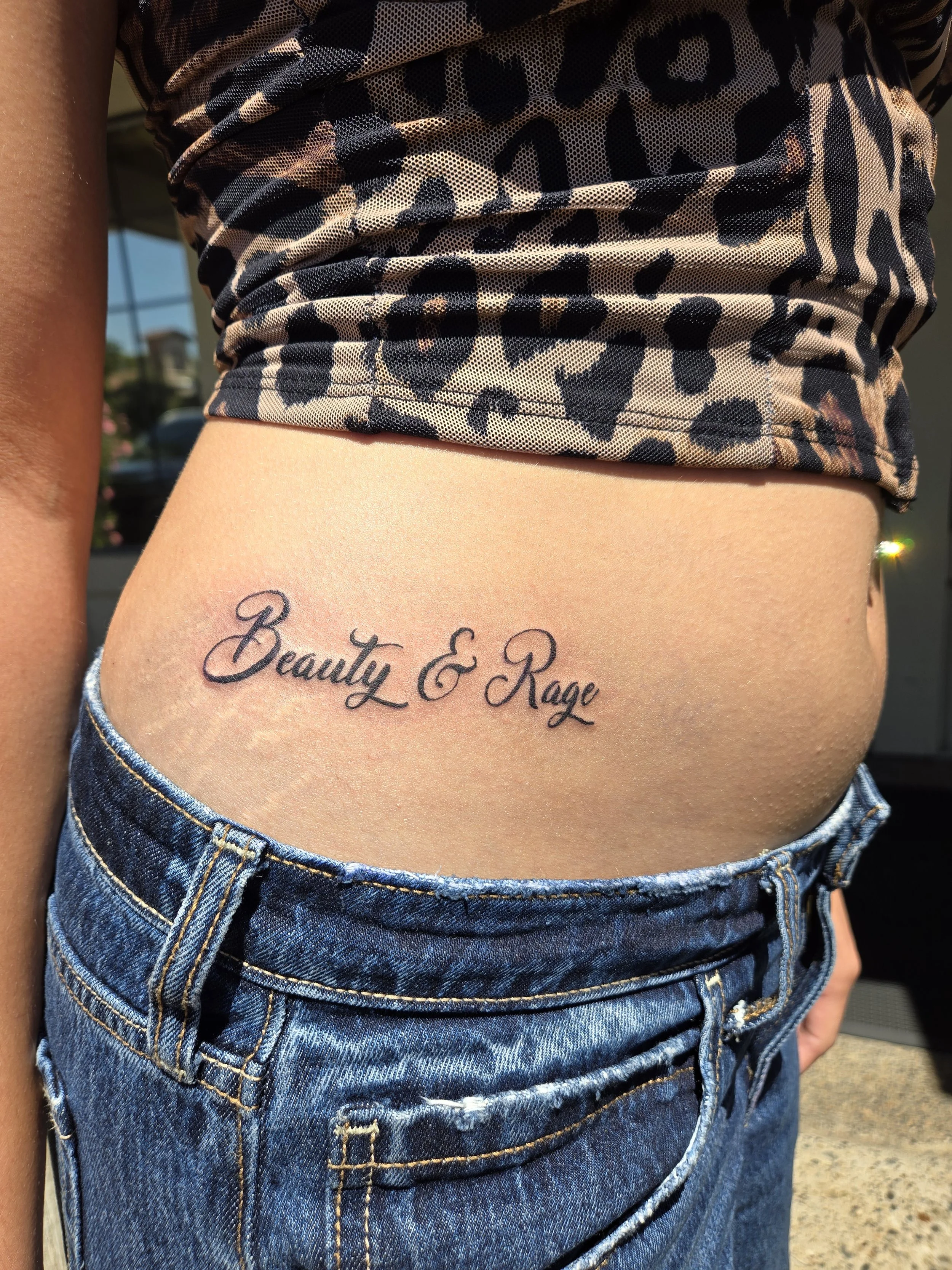 A close-up of a person's midsection showing a tattoo that reads 'Beauty & Rage' on the lower ribs, with the person wearing a leopard-print top and denim shorts.