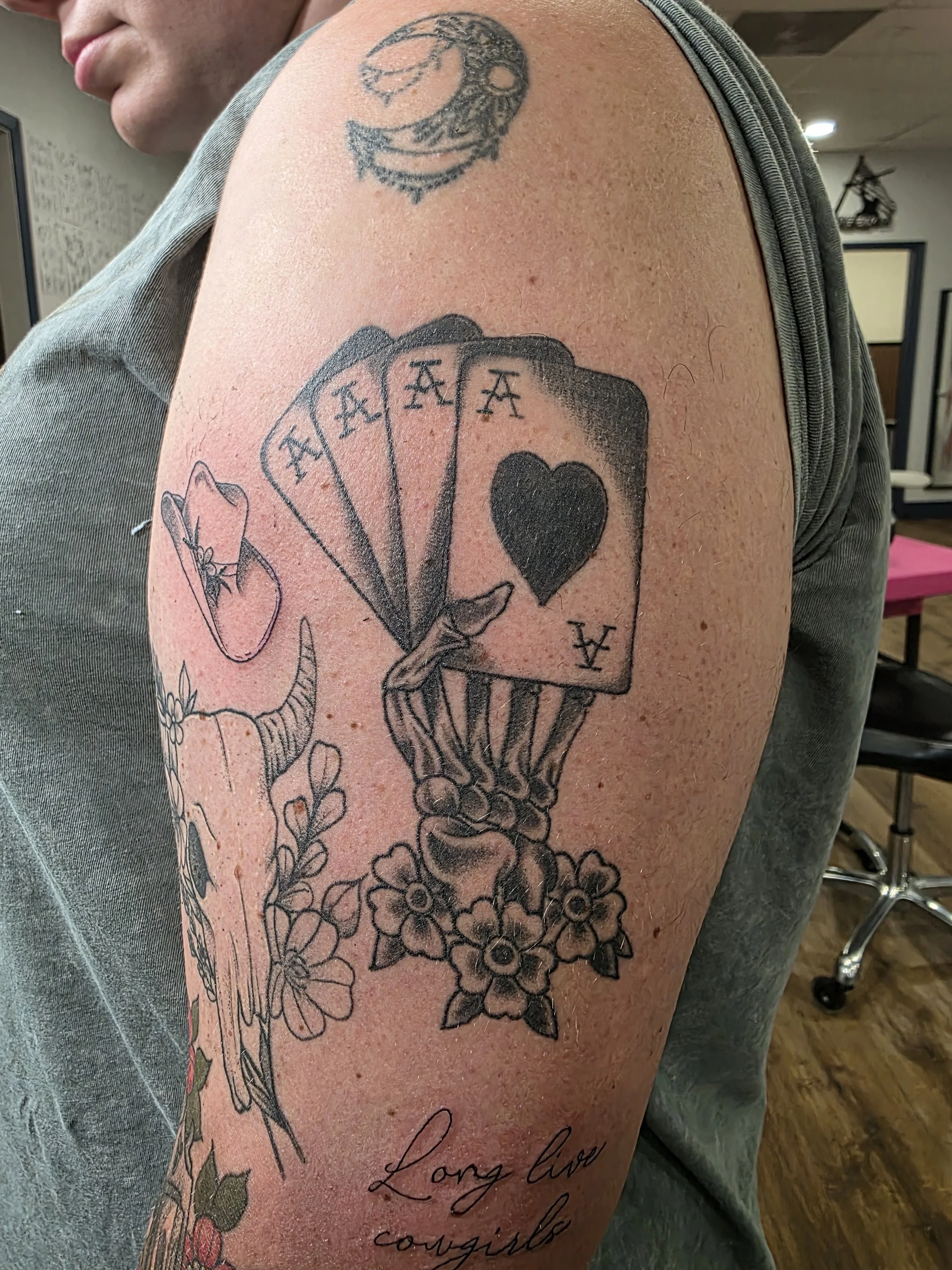 A person's upper arm with tattoos, including a black heart, a fan of playing cards, a snake, and a floral design, along with cursive words.