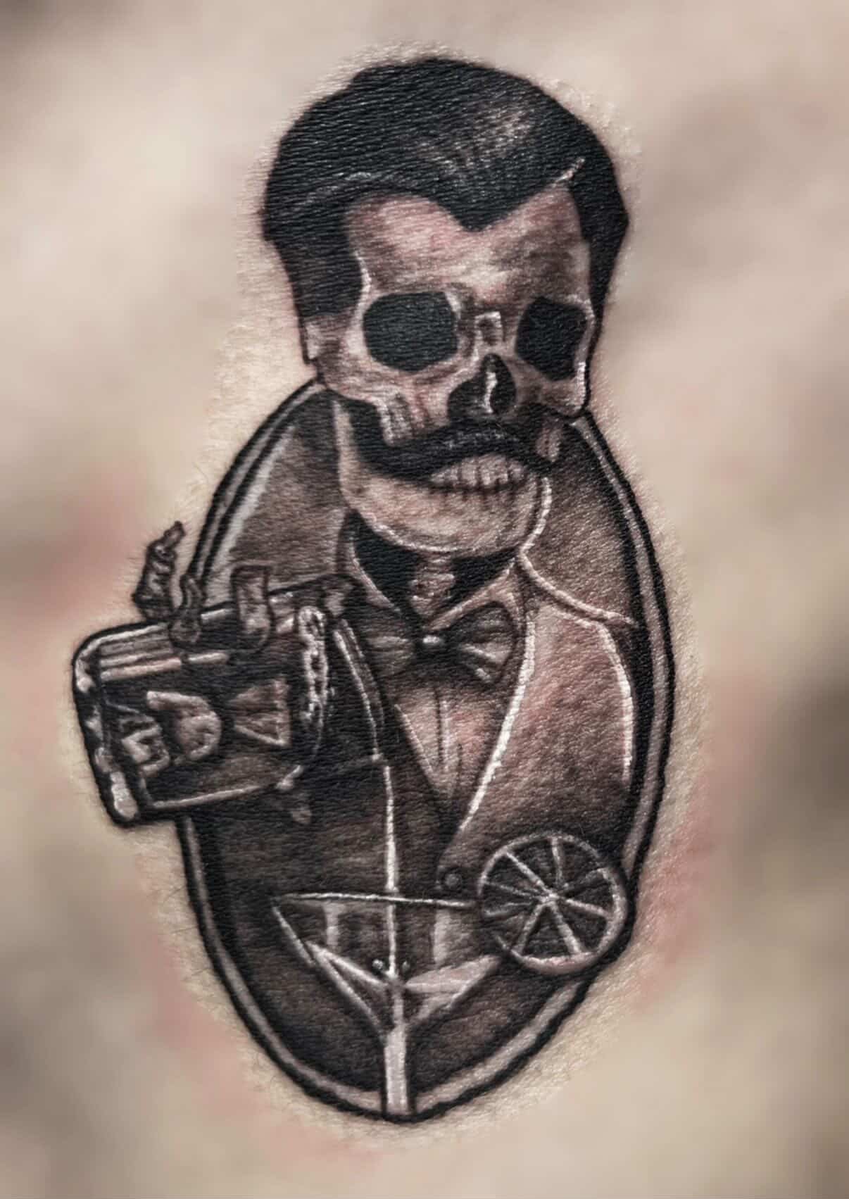 Tattoo of a skeleton wearing a tuxedo, pointing with one finger, inside a martini glass shape.