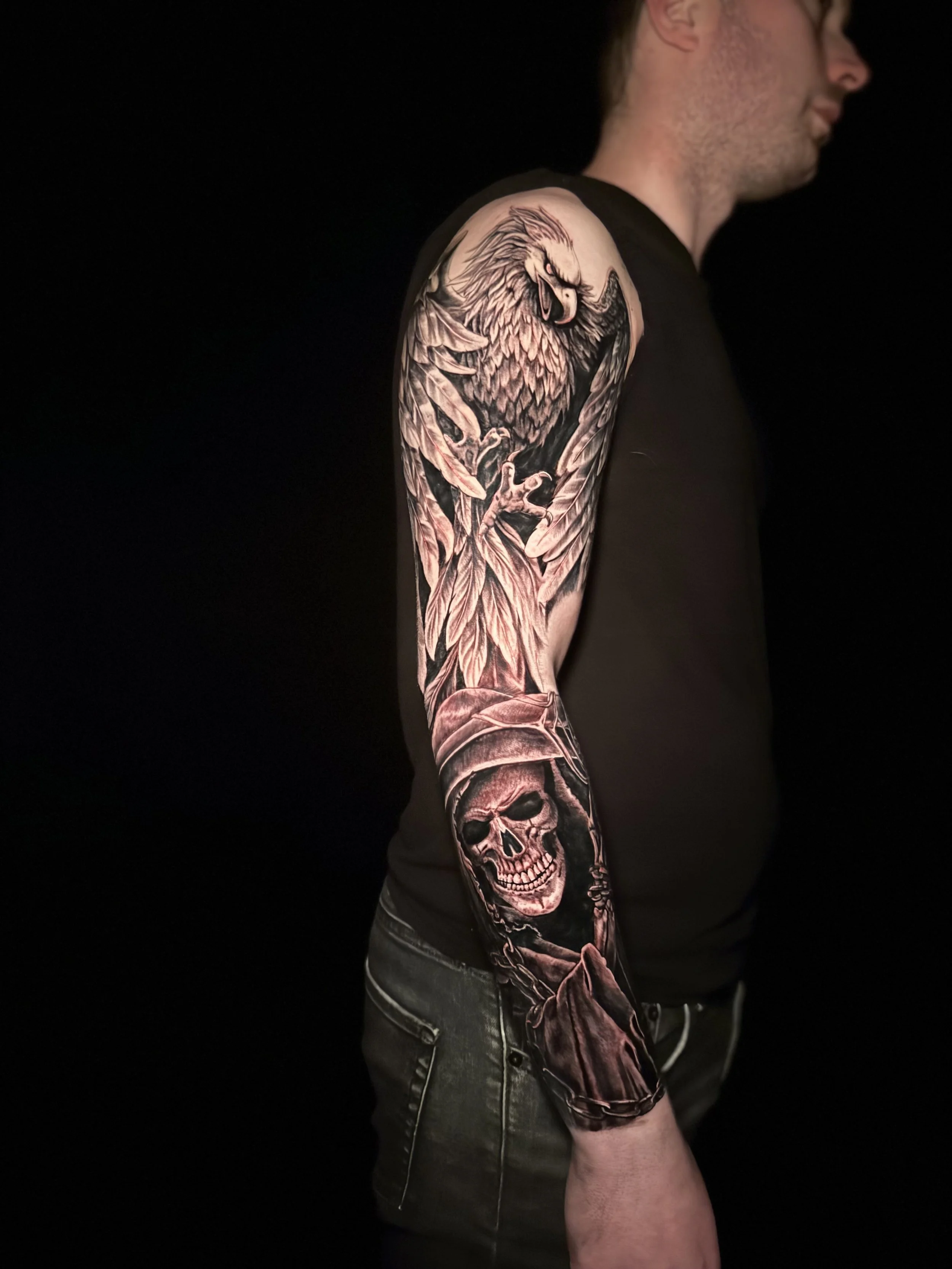 A person with a tattoo of an eagle, wings, and skulls on their arm, wearing a black shirt and denim shorts, with a dark background.
