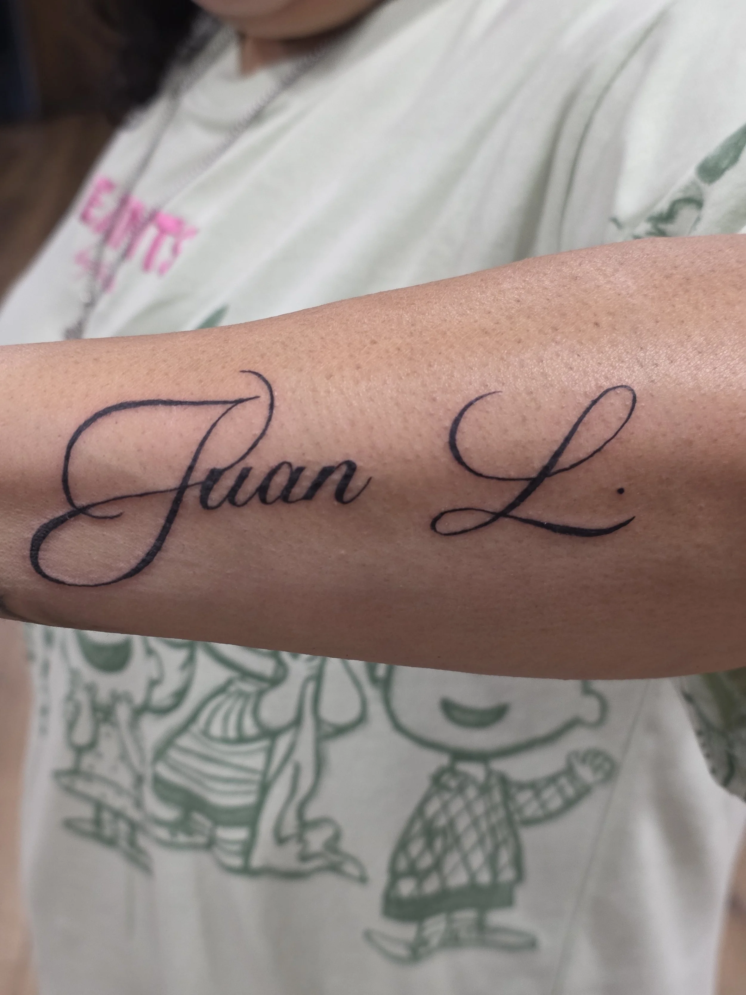 Close-up of a person's forearm with a tattoo that reads 'True L' in black cursive script, with part of the tattoo visible.