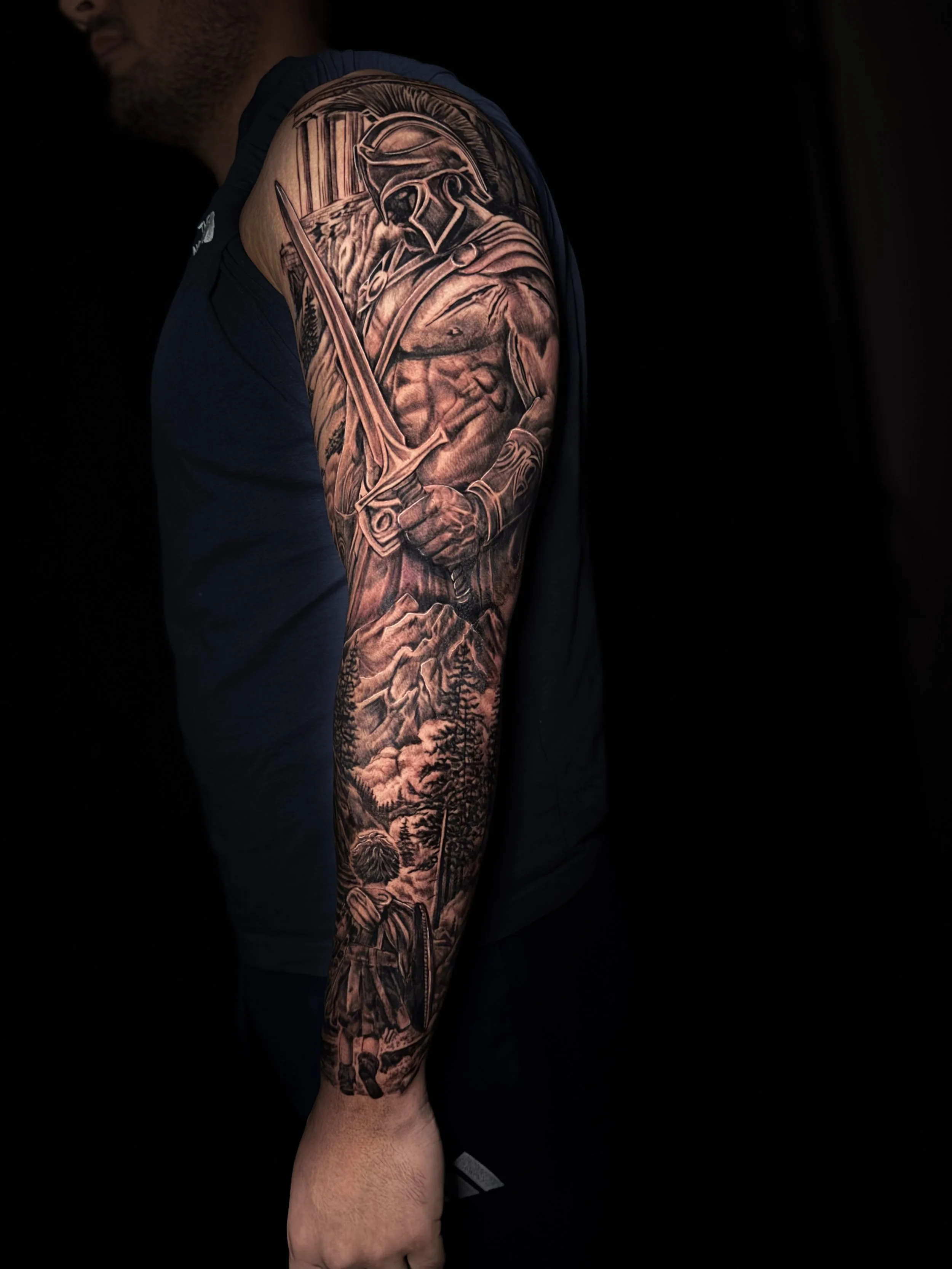 A person with a detailed black and gray tattoo sleeve featuring a warrior with a helmet, a sword, and forest scenery on their arm.
