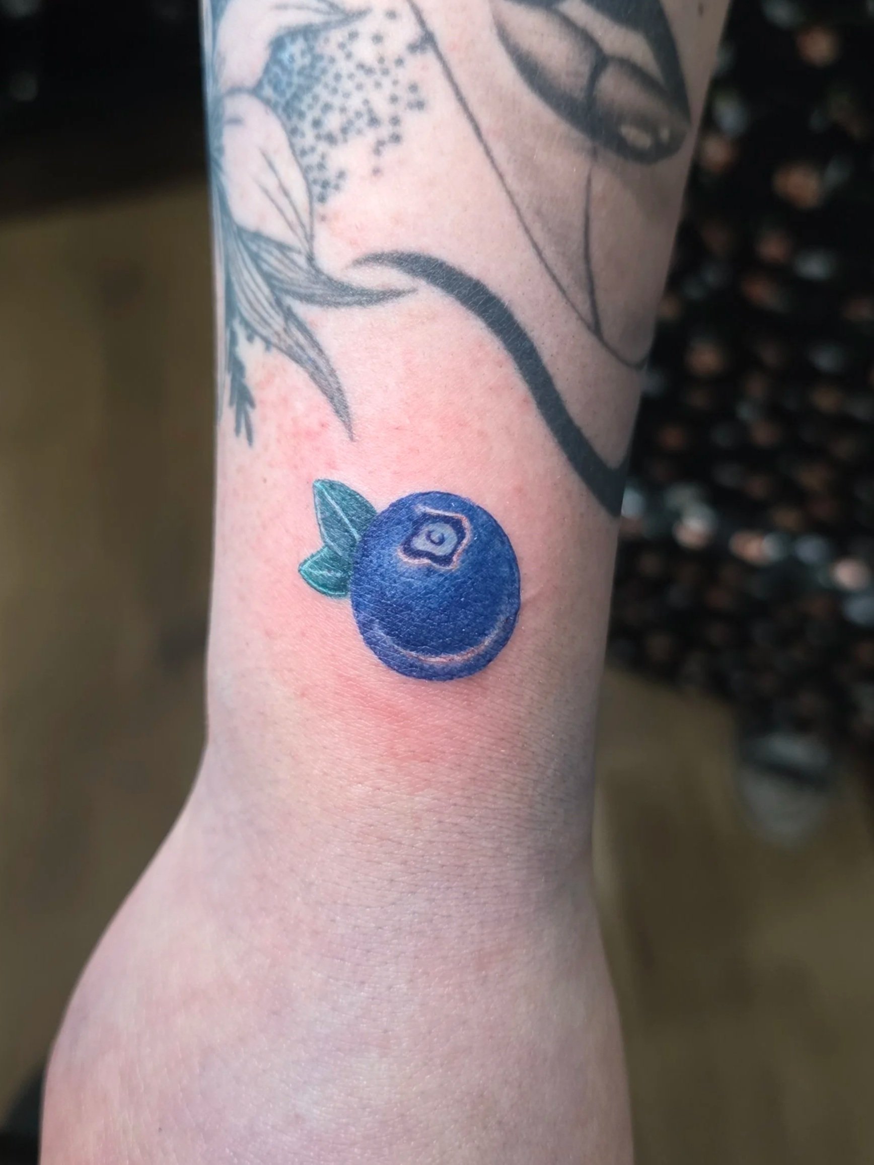 Tattoo of a blue berry with green leaves on person's arm.