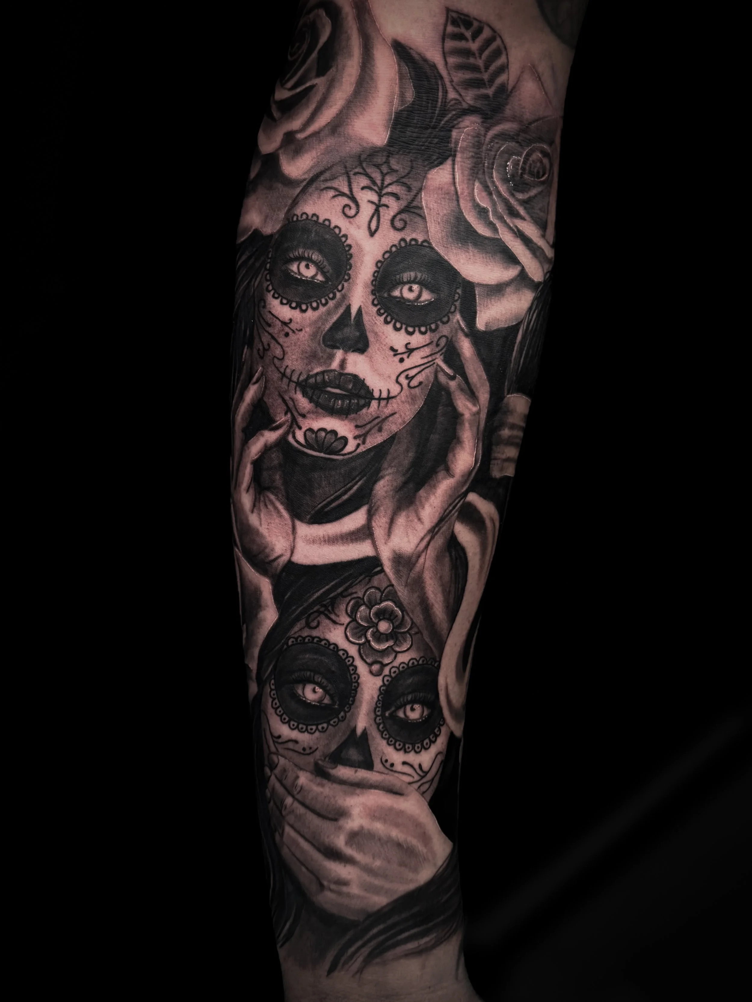 Tattoo of a woman with a sugar skull face design, featuring large, detailed eyes, dark lipstick, and decorative patterns, on an arm.