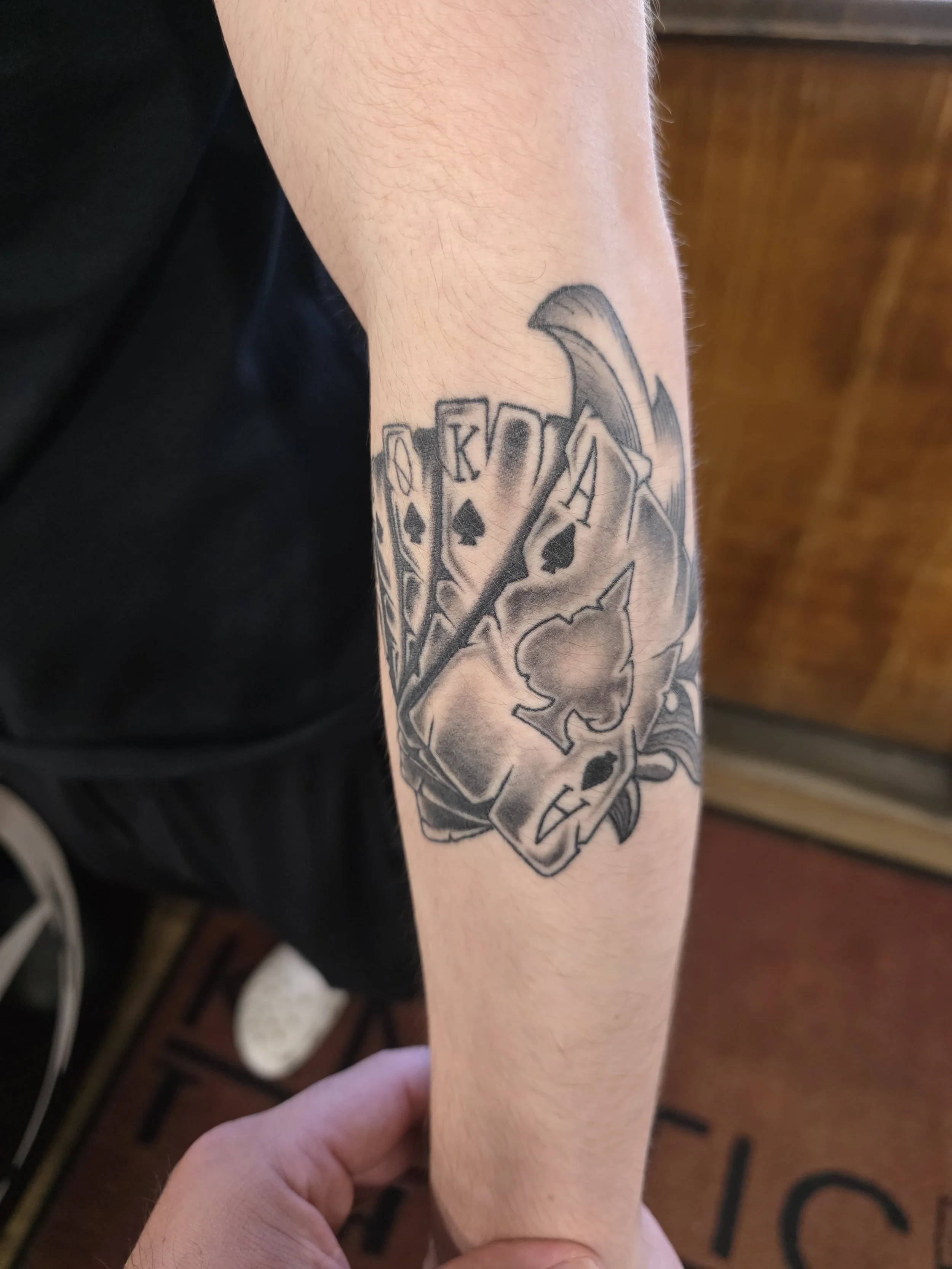 Tattoo on person's forearm featuring playing cards spelling 'KOTA', a stylized wing, and a flower.