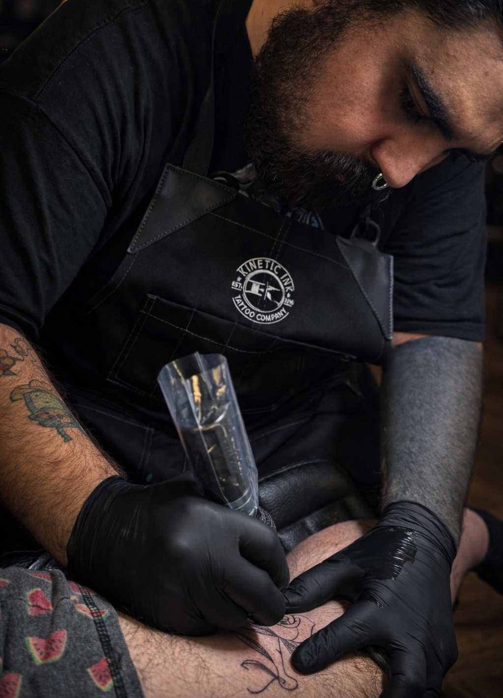 A tattoo artist with a beard wearing a black apron and black gloves, tattooing a design on a person's leg.