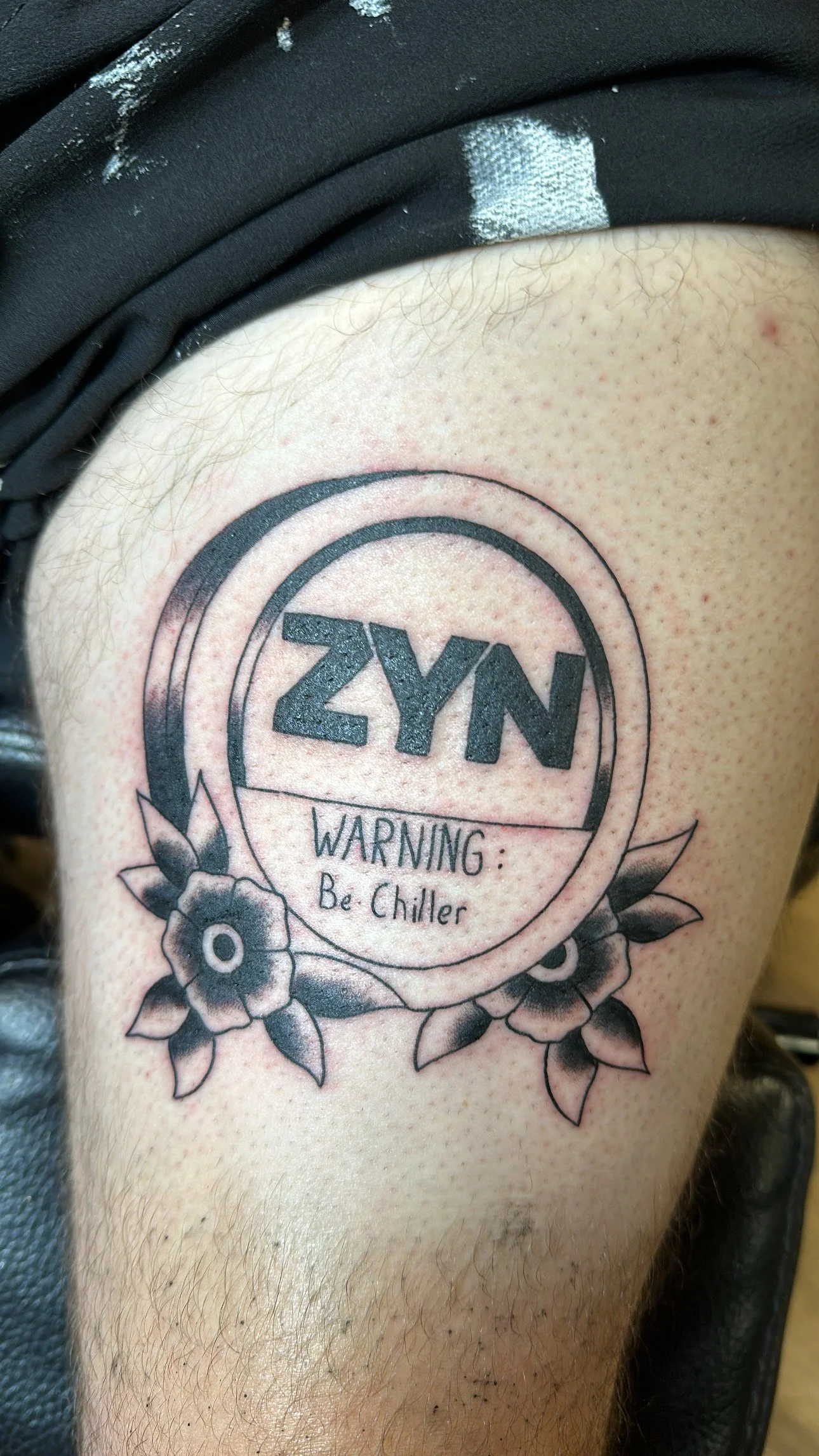 Tattoo on person's upper arm with the word 'NY' in bold, surrounded by a circle, and a banner below reading 'WARNING: Be Chiller' with floral design.