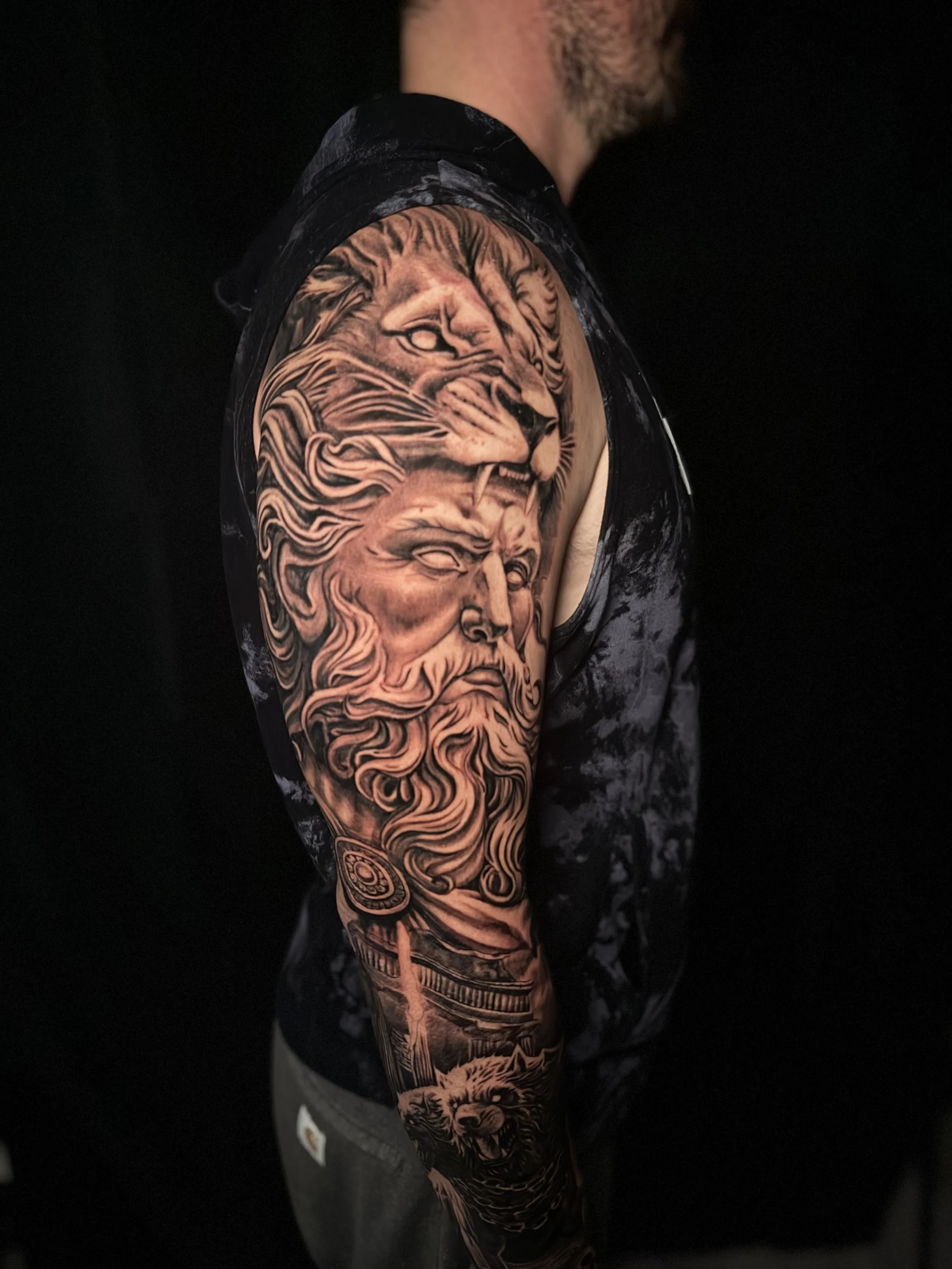 A person's arm with a detailed black and gray tattoo of a lion and a bearded man on the upper arm and shoulder, wearing a black sleeveless shirt against a dark background.
