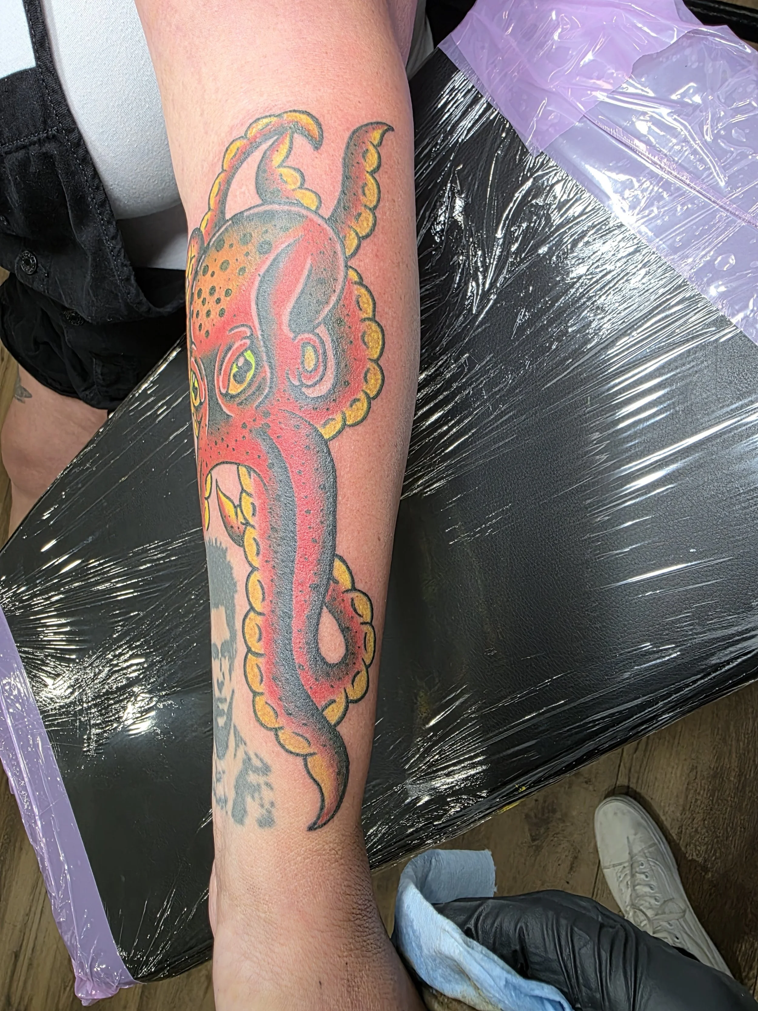 A person's forearm with a colorful tattoo of a squid on the skin, sitting on a black surface covered with clear plastic wrap, with a foot visible in white shoe in the lower right corner.