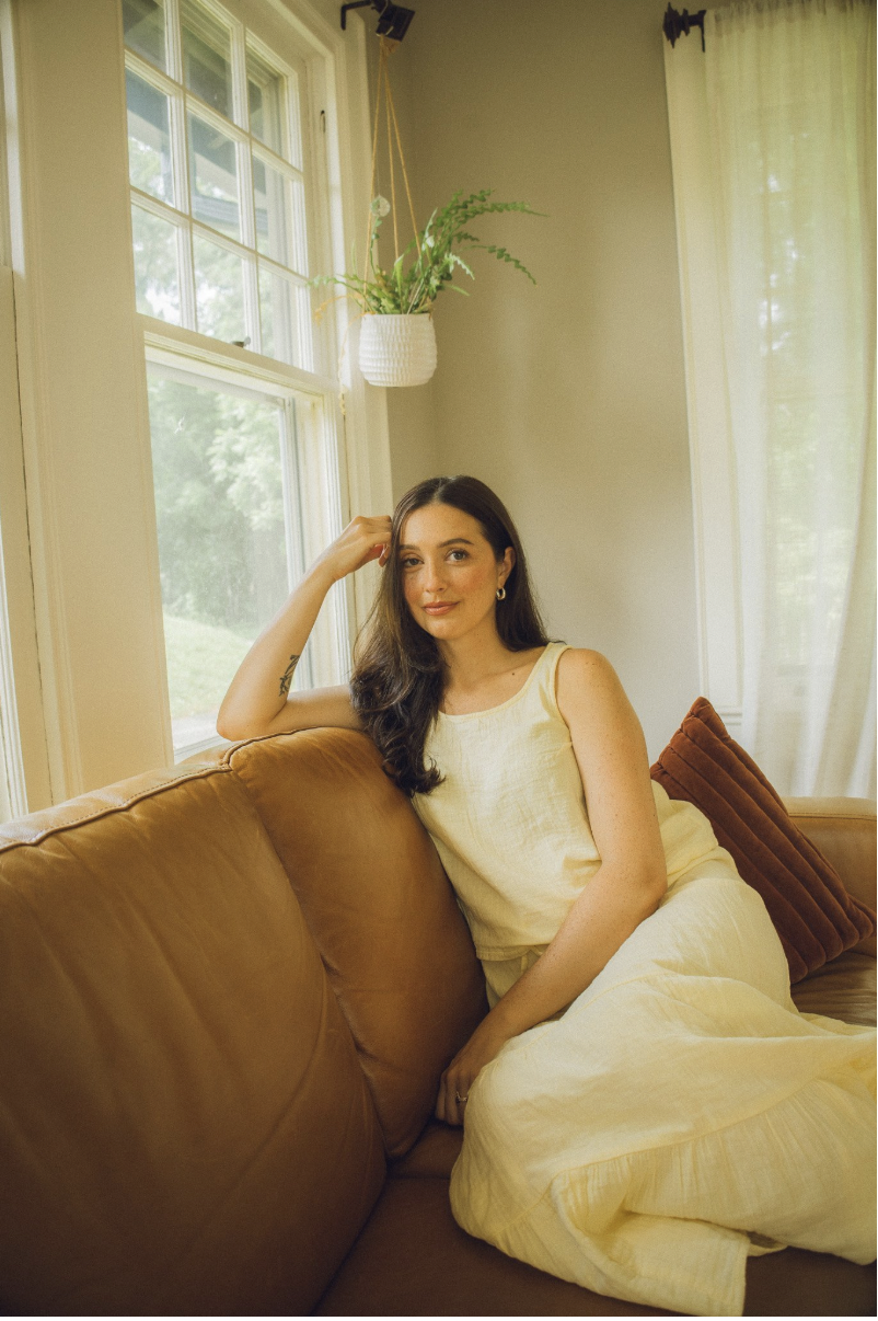 A woman with dark hair in a yellow dress sitting on a tan leather couch near windows with white curtains and a hanging potted plant.