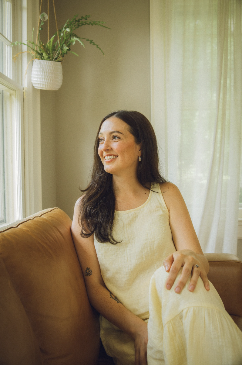A woman with long, dark hair sitting on a beige sofa near a window with sheer cream curtains, smiling and looking to her left. There is a hanging white planter with green plants in the corner of the room.