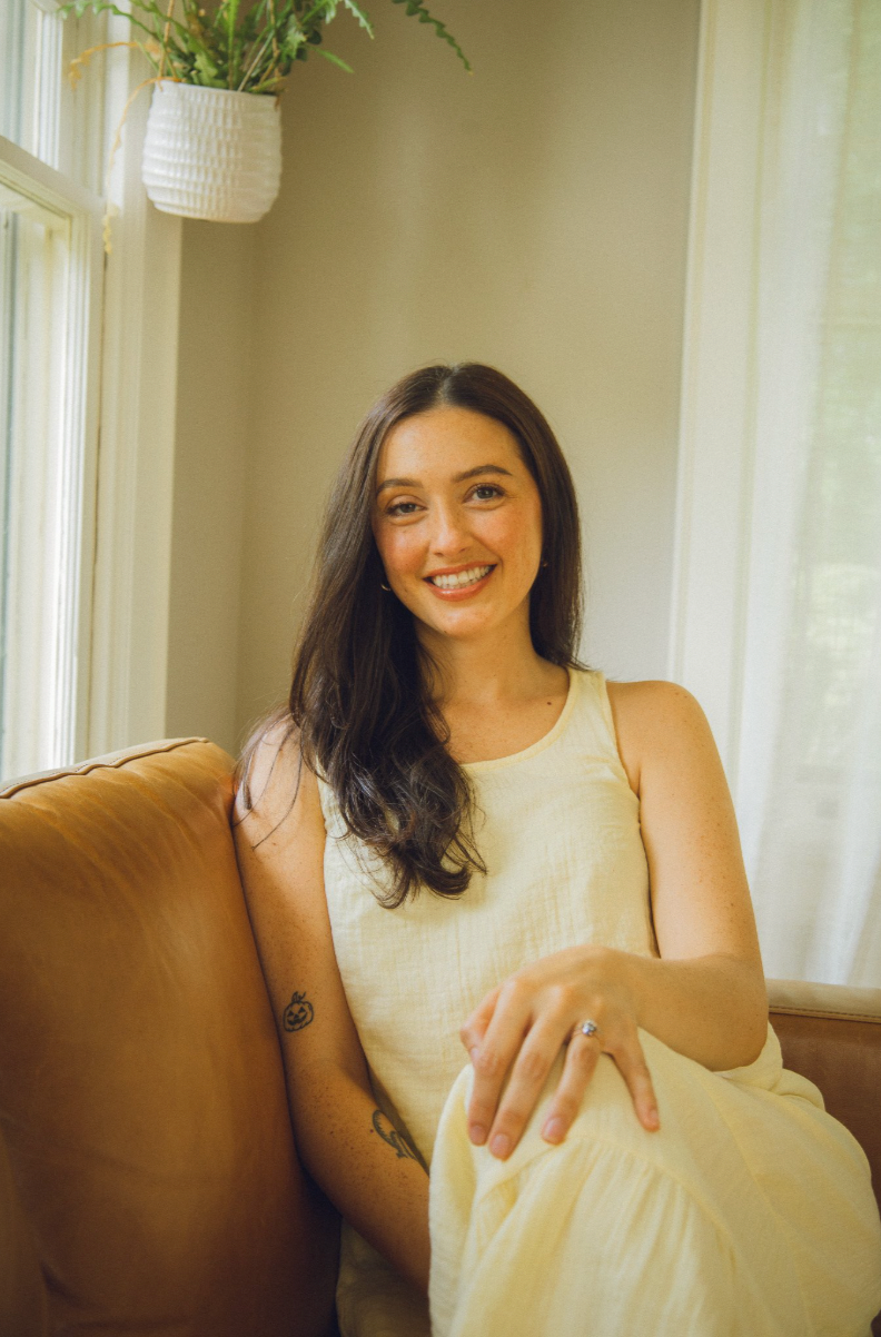 A smiling woman with long dark hair sitting on a beige couch in a well-lit room near a window with sheer white curtains and a hanging plant.
