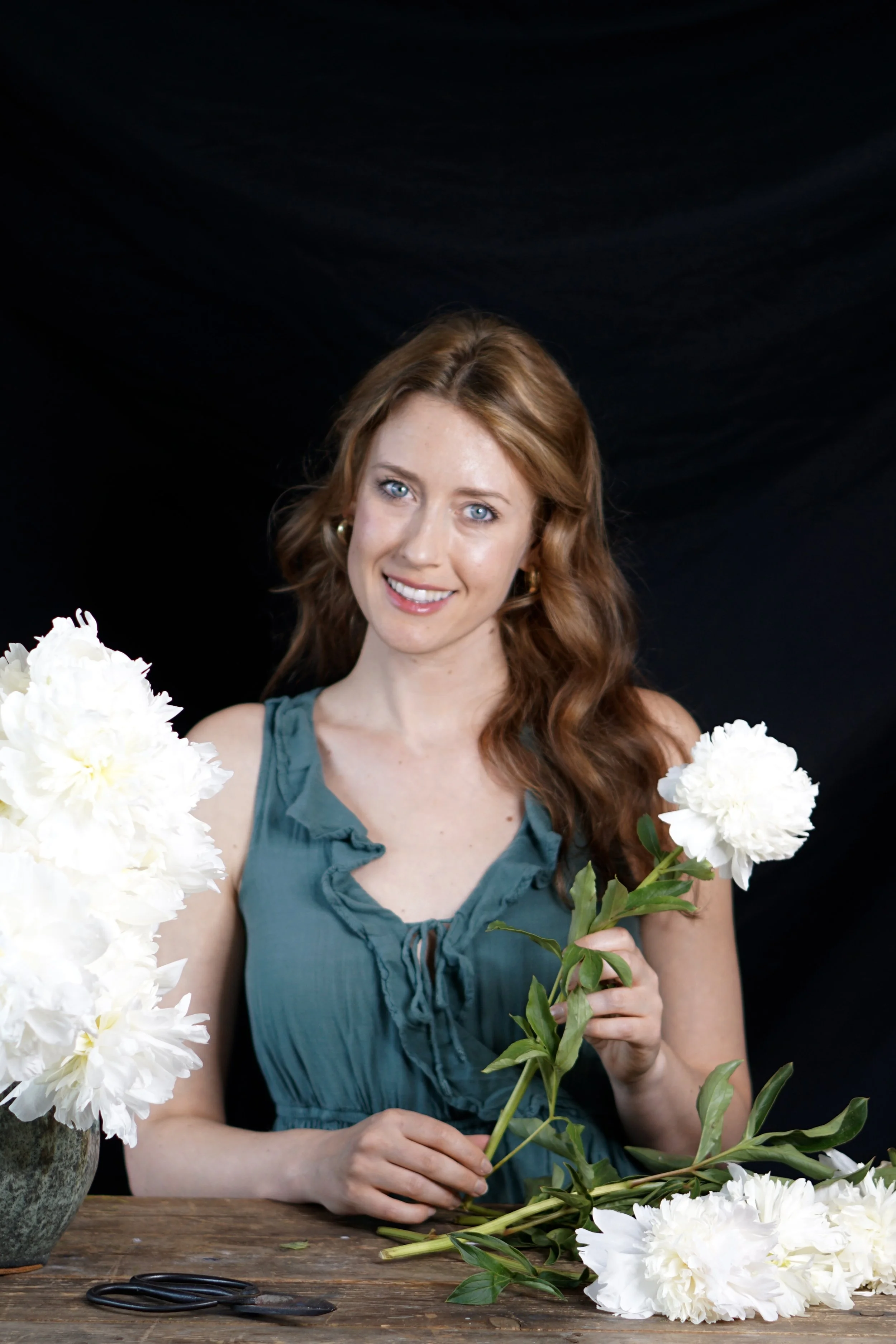 A woman with red hair and blue eyes is smiling while holding a white flower. She is wearing a teal ruffled dress and is surrounded by white flowers on a wooden table.
