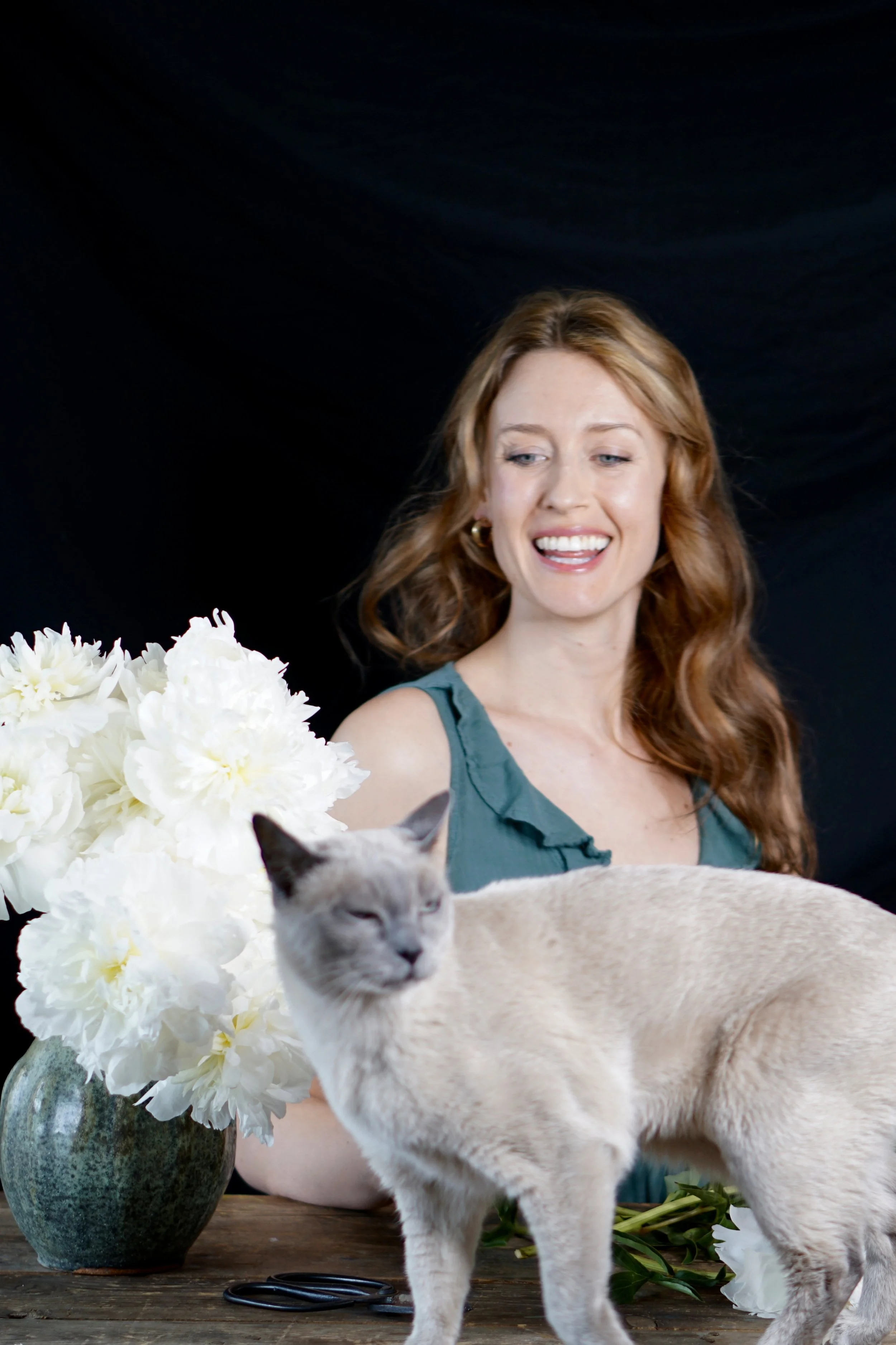 A woman with long red hair wearing a sleeveless teal top, smiling, with a large bouquet of white peonies in a green vase on a wooden table, a Siamese cat in the foreground, and a dark backdrop.