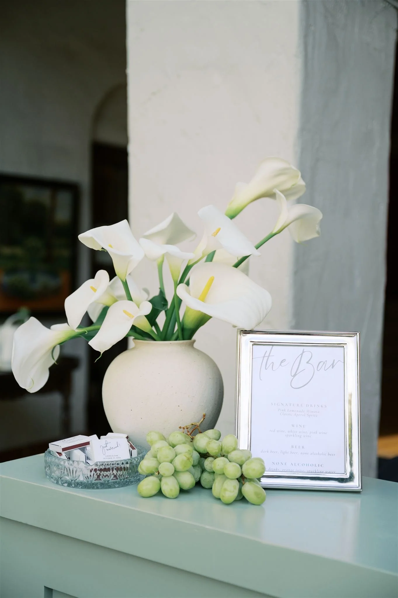 A white ceramic vase with white calla lilies, a framed menu for signature drinks, grapes, and a business card holder on a teal table