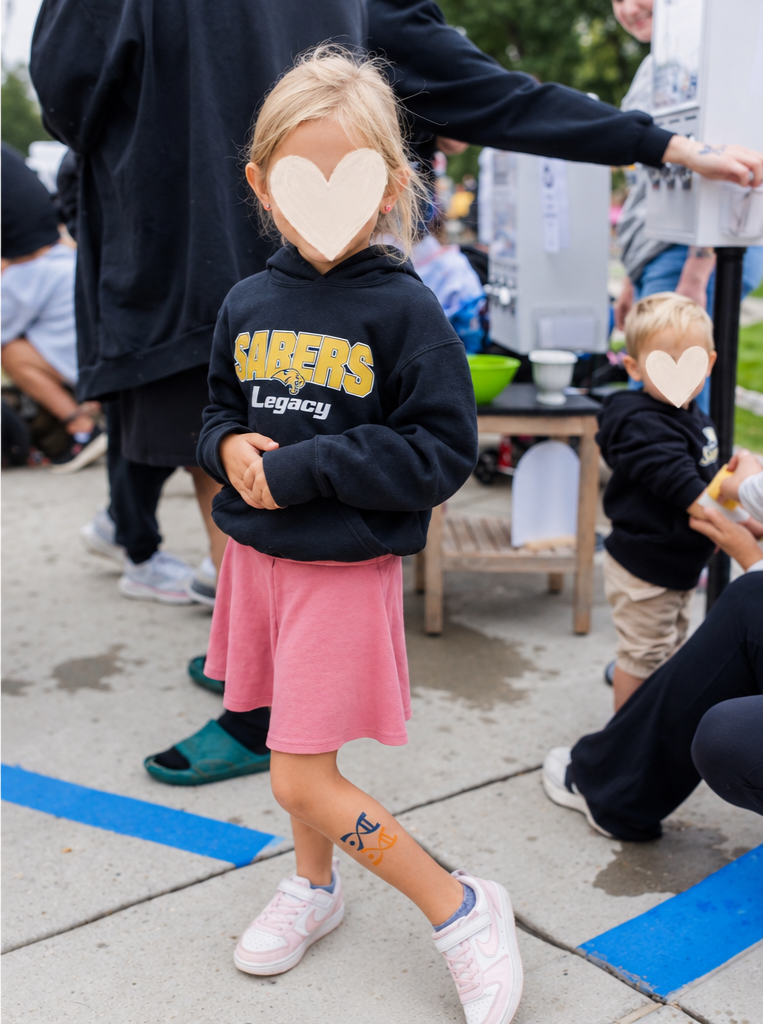 Child with a face covered by a heart-shaped mark, wearing a Sabers Legacy hoodie, pink skirt, and pink sneakers, standing outdoors during an event.