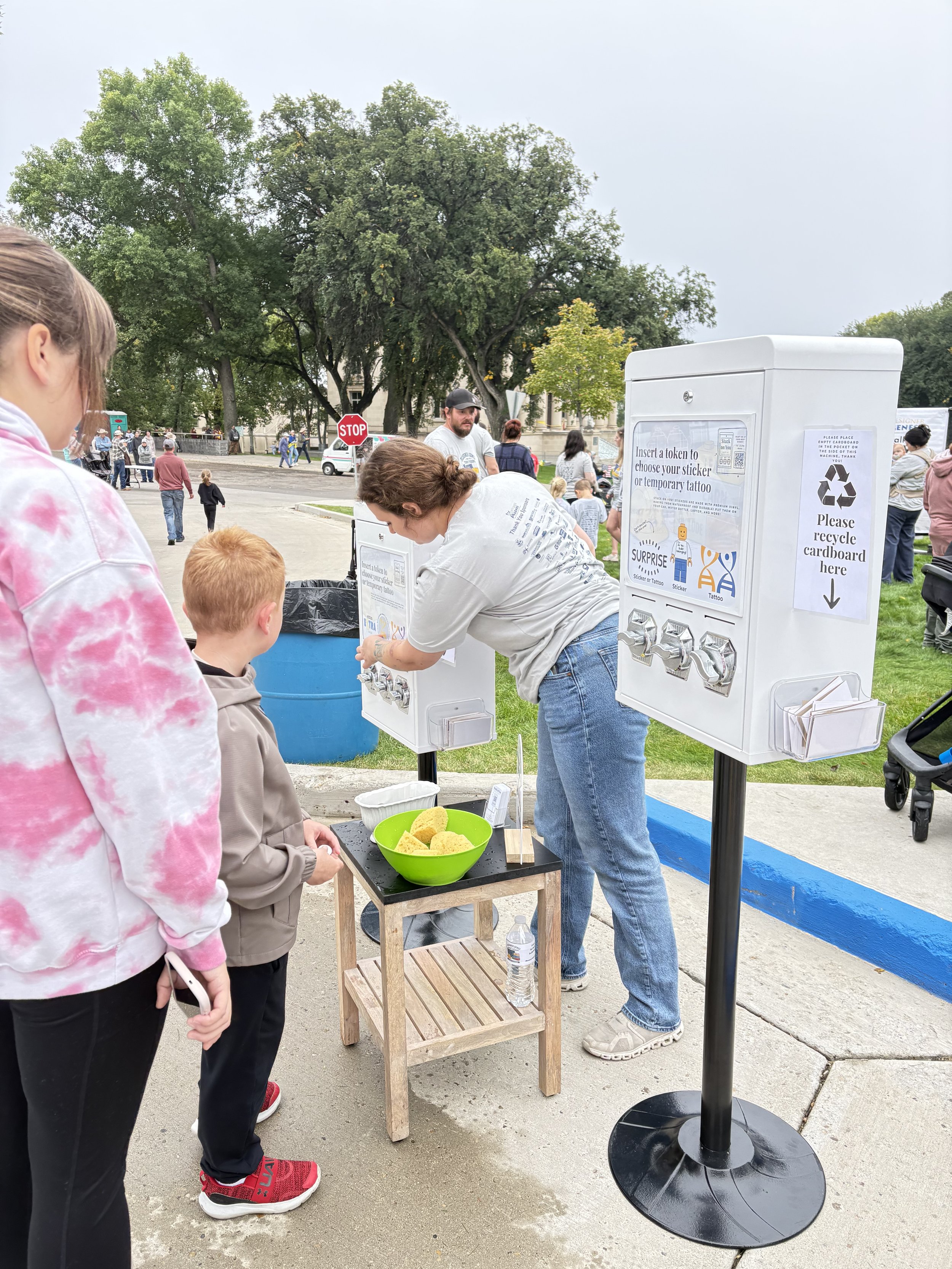 People at an outdoor event in a park, with a ticket or sticker vending station and a waste bin nearby. A child and an adult are selecting tokens or stickers from the station.