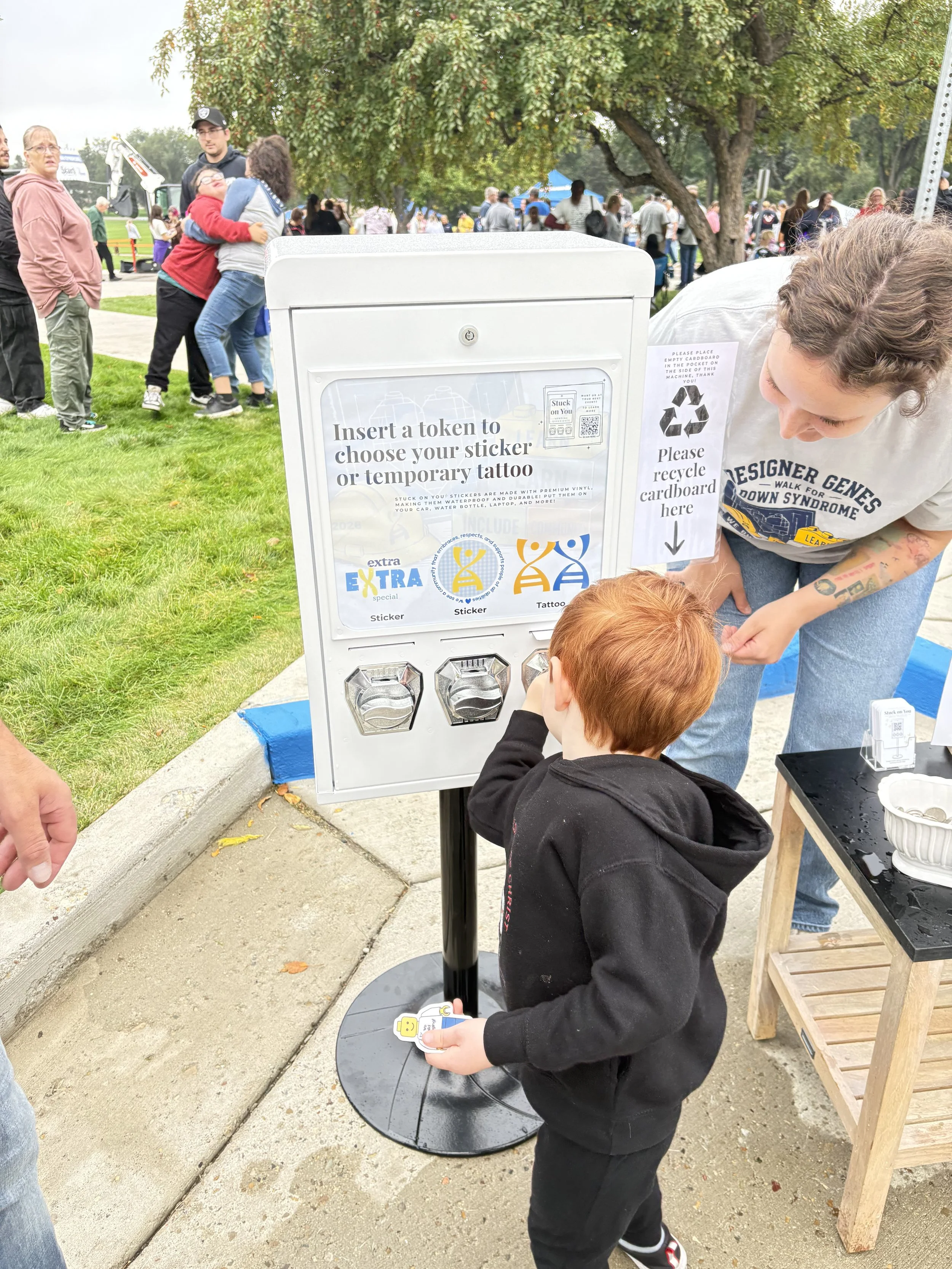 A boy with red hair holding a sticker with a Lego character in front of a donation bin at an outdoor event, with people and trees in the background.