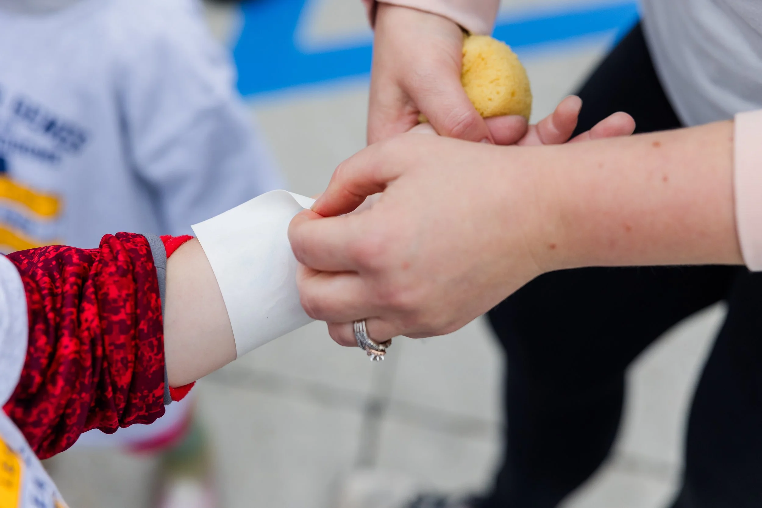 A person holding a yellow sponge, with a child's arm in the foreground. The child is wearing a red patterned sleeve. The background shows a person in a light-colored shirt and another in dark clothing.