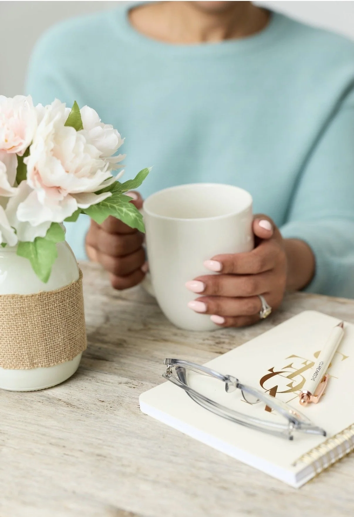 A person with medium to dark skin tone wearing a light blue top holding a white mug, sitting at a table with a white notebook, a pair of glasses, a pen, and a vase with pink and white flowers, in a cozy setting.