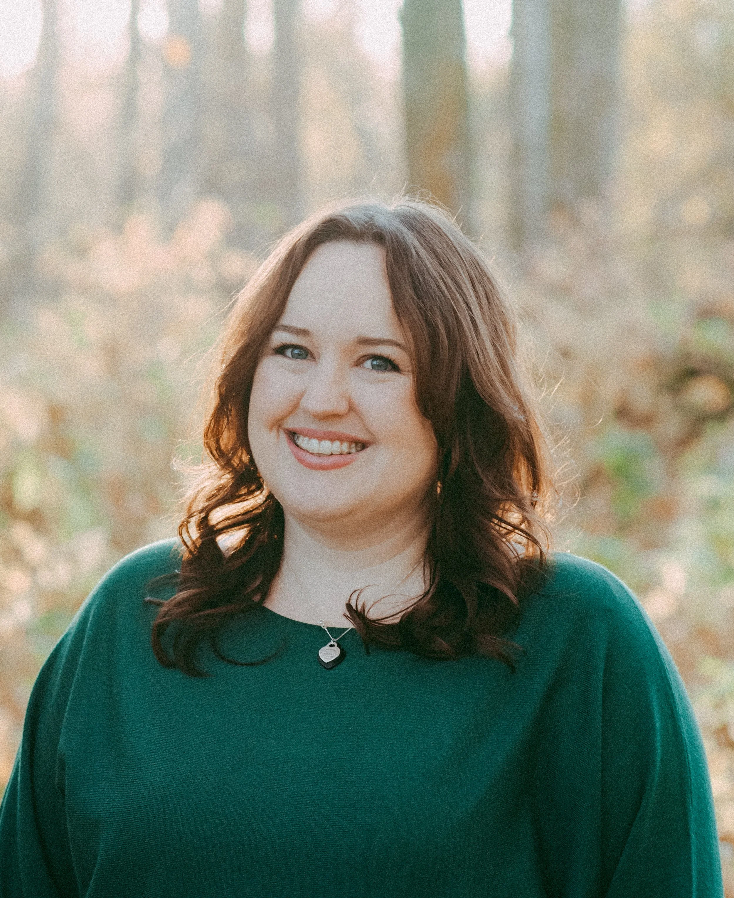 Close-up of a smiling woman with blue eyes and long wavy brown hair, wearing a dark green top and a silver necklace with a circular pendant, standing outdoors with blurred trees and autumn leaves in the background.