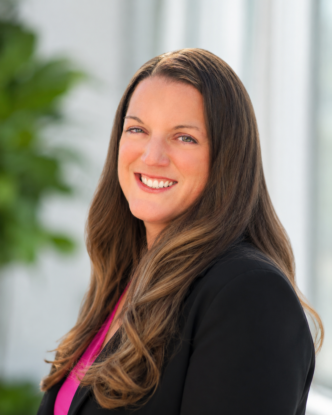 Close-up portrait of a woman with long brown hair, smiling with a slight squint in her eyes, standing in front of a blurred background with natural light.