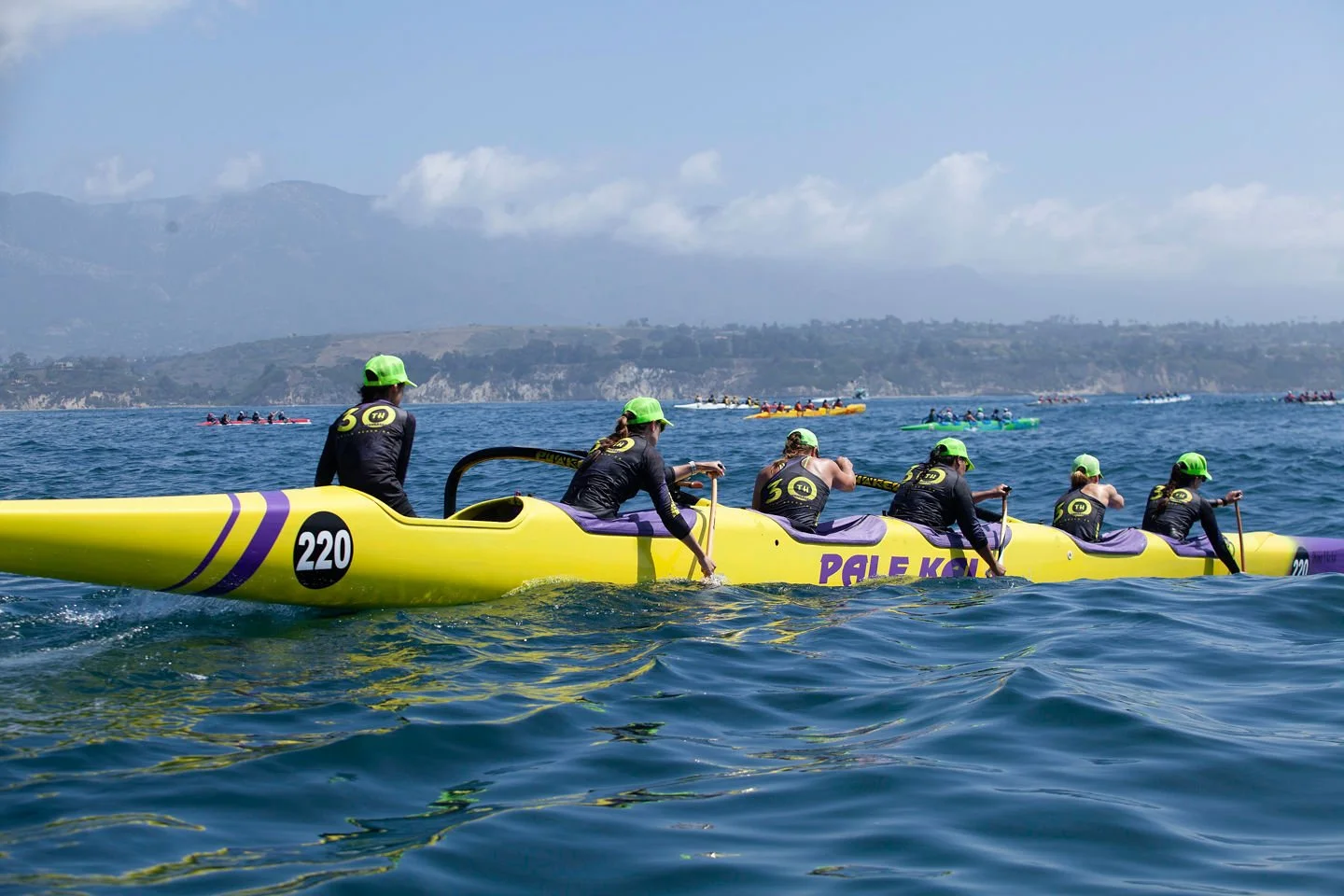 A team of seven rowers in a yellow canoe with purple accents and a black circle with the number 220, paddling on the water with other boats in the background and mountains under a partly cloudy sky.
