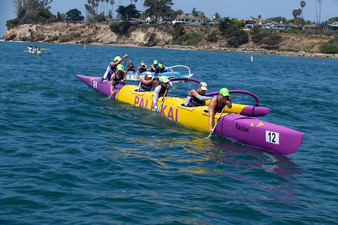 A group of people paddling a colorful dragon boat on the water during a race near the coast with houses and trees on the shoreline.