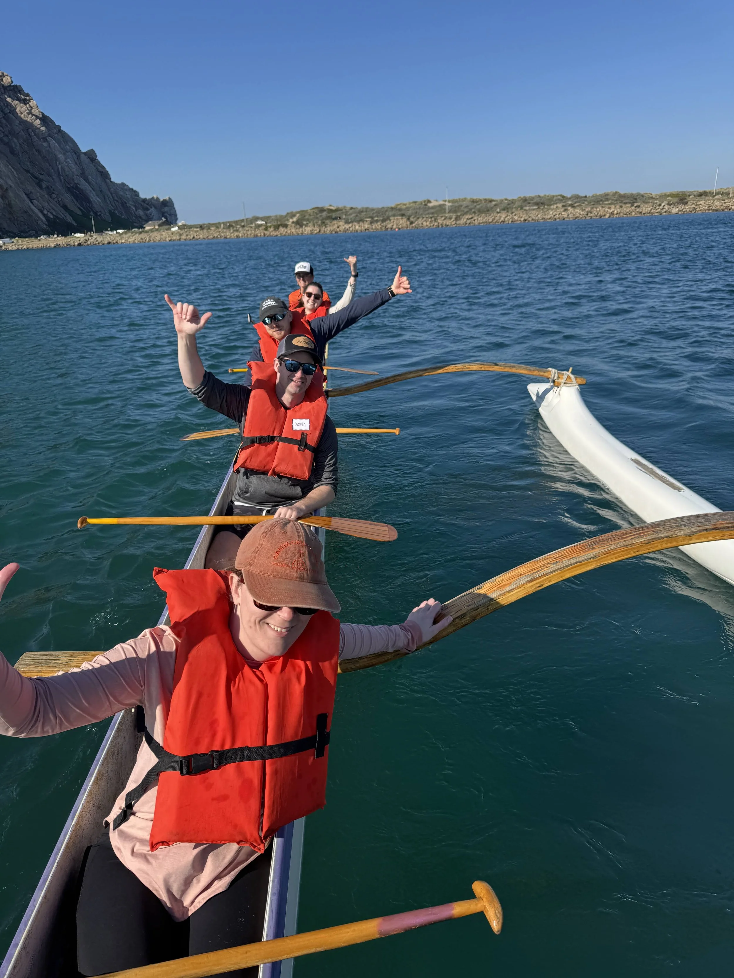 Group of people in a boat on the water, smiling and making hand gestures, with a rocky coastline and clear blue sky in the background.
