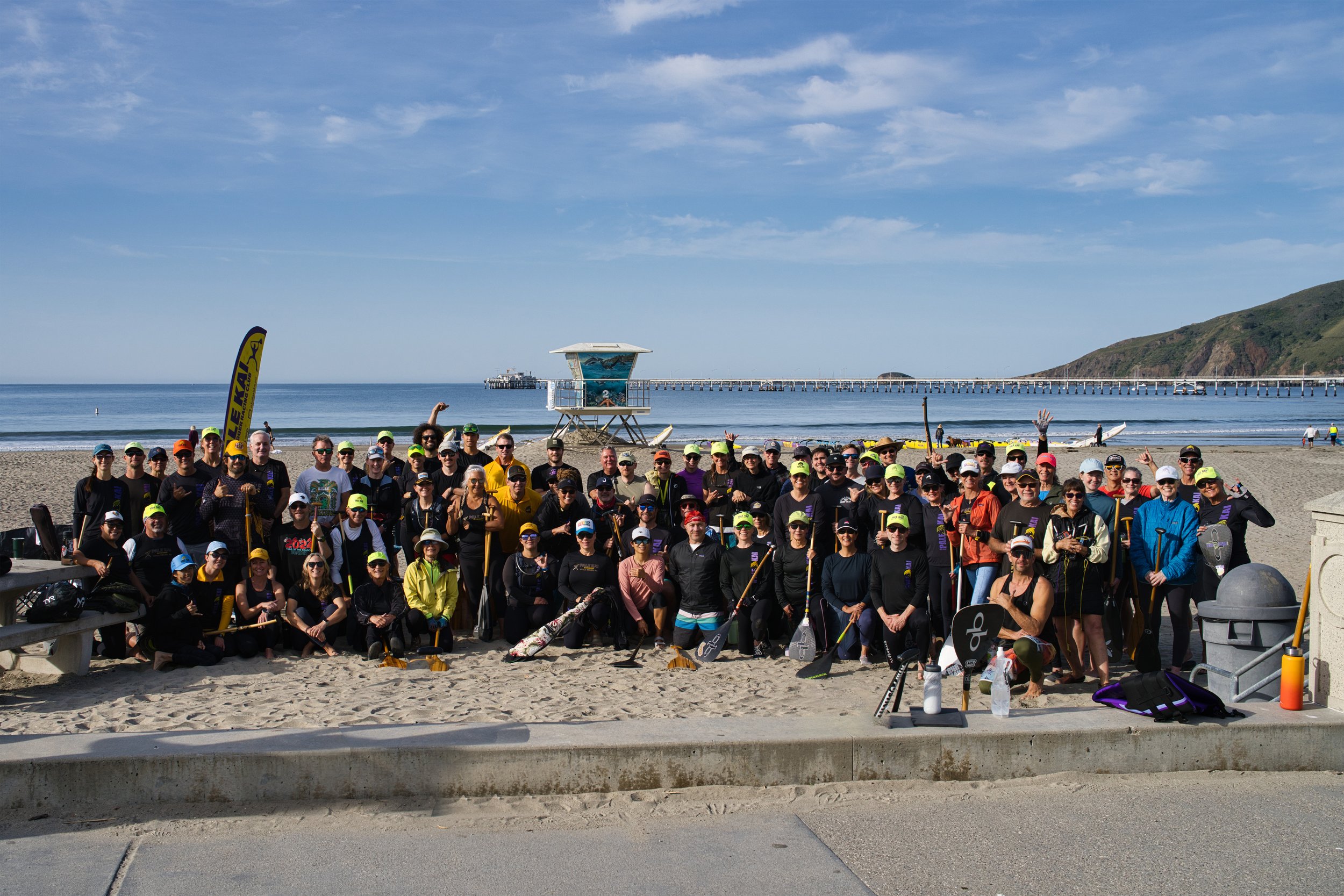 A large group of people gathered on a sandy beach, posing for a group photo. They are wearing casual clothes, hats, and sunglasses. In the background, there is a pier extending into the ocean, with a lifeguard tower near the shoreline, and a hill on 