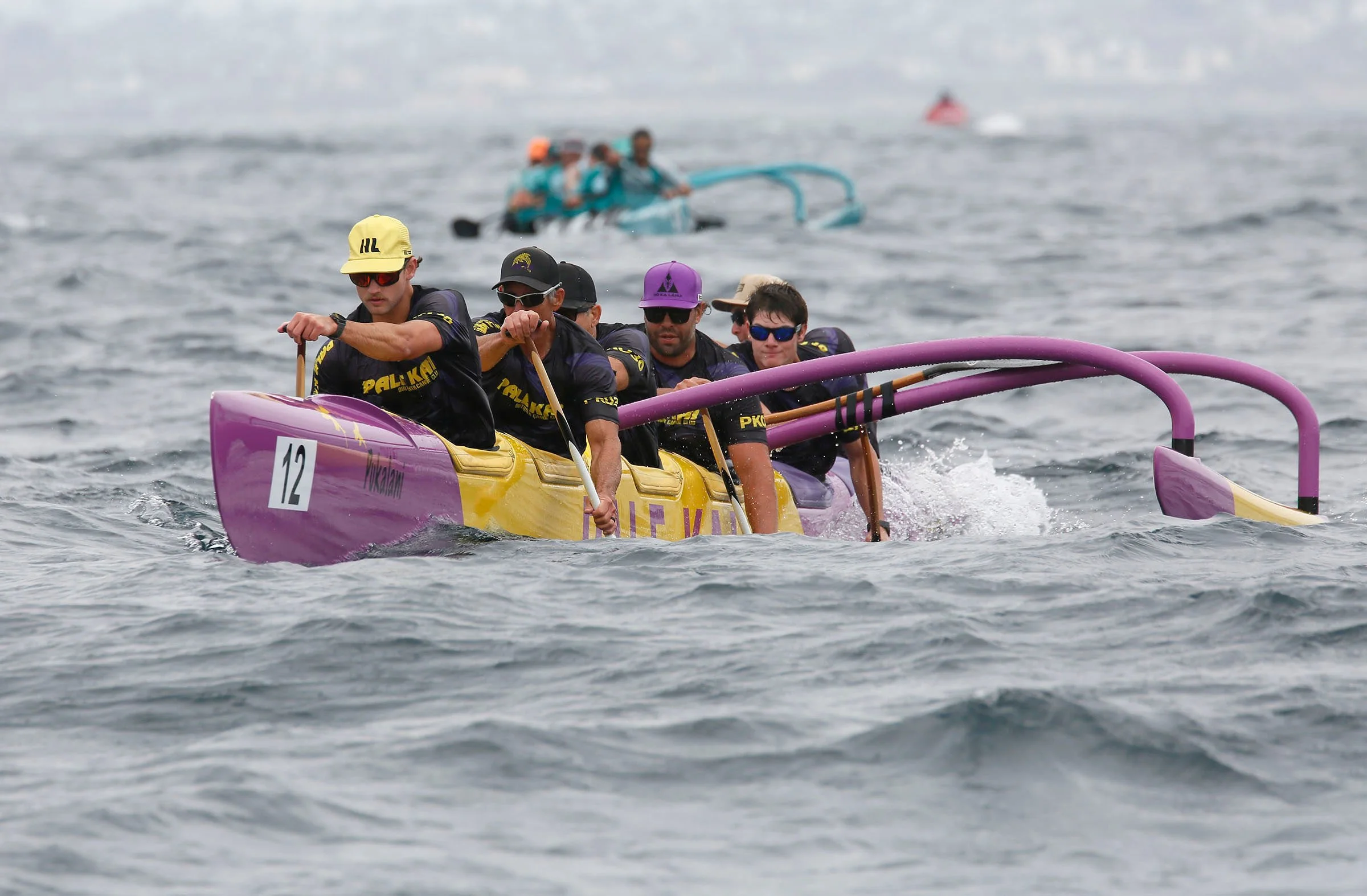 A team of five people participating in a boat race on the water, with some in the foreground and others in the background, paddling a purple and yellow boat.