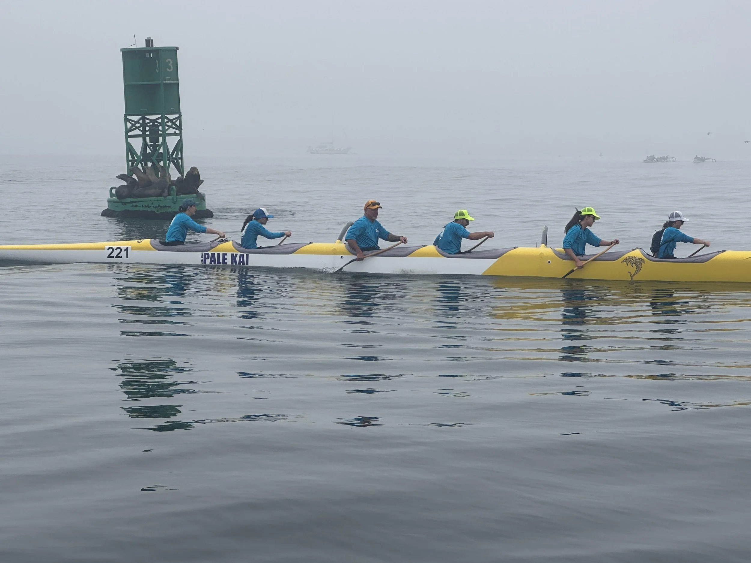 A group of six people wearing blue shirts and yellow or white hats paddling a yellow canoe named 'Pale Kai' in foggy water. A green buoy with the number 33 and a sea lion statue is visible in the background, along with boats on the horizon.