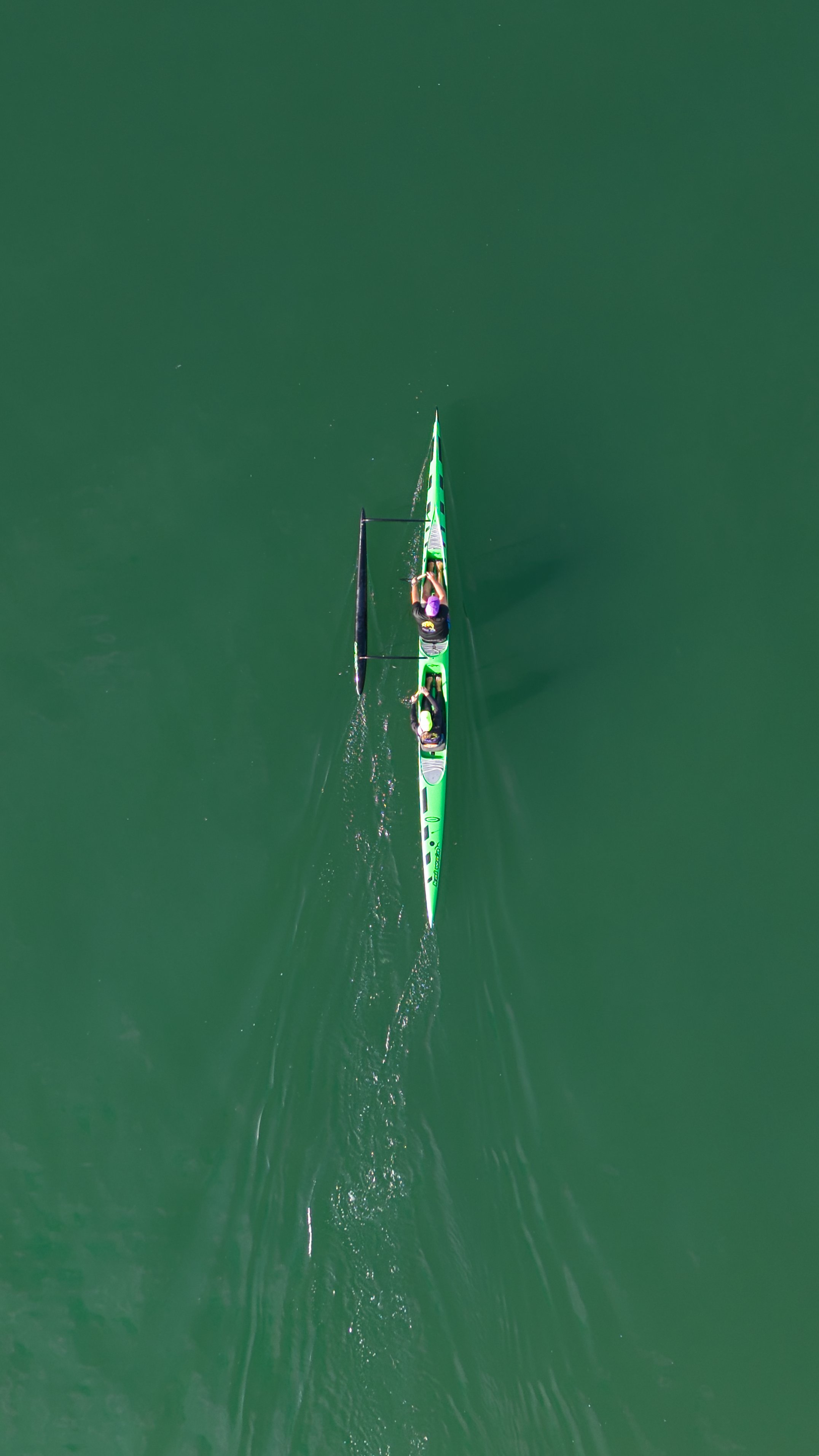 An aerial view of two people rowing a green racing shell on calm green water.