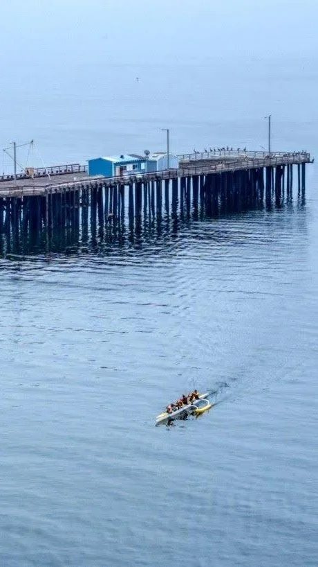 A long wooden pier extending into calm water with small blue building at the end, and a group of people paddle boating nearby.