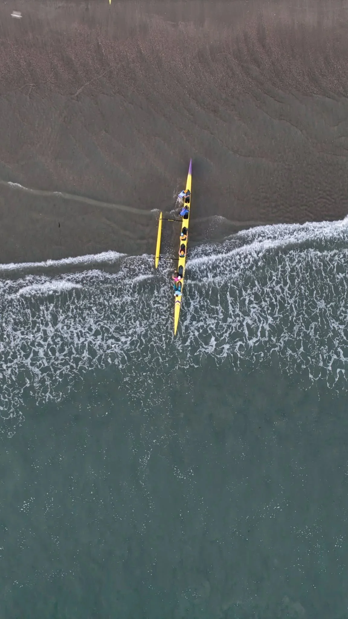 An aerial view of a yellow boat with five people on board, riding near the shoreline where waves are washing onto the sand.