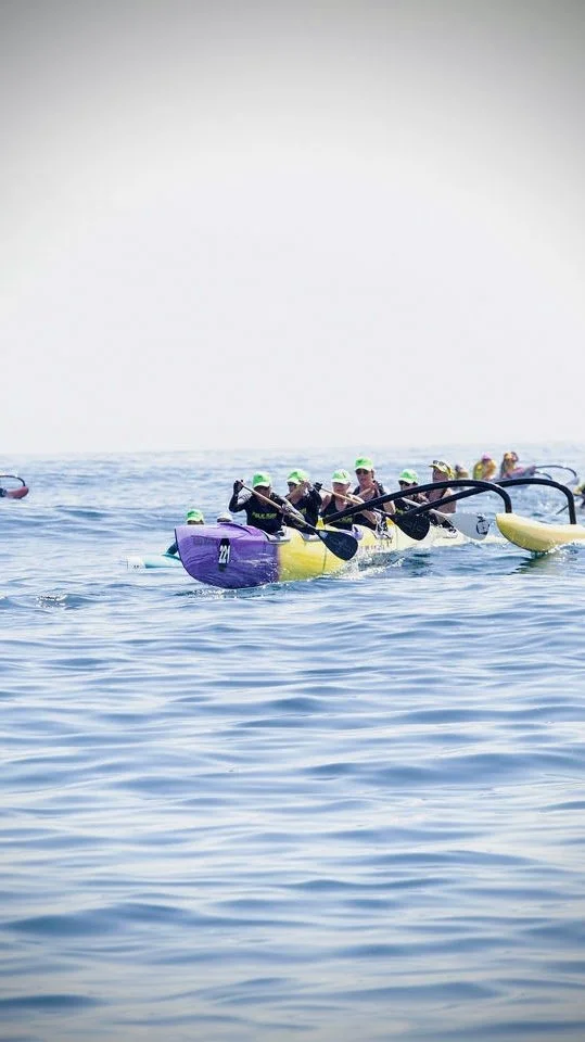 Group of people paddling a dragon boat on open water, wearing life jackets and hats.