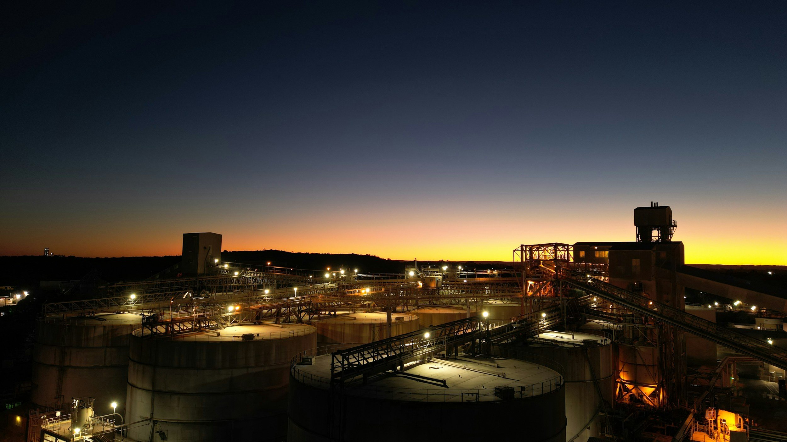 Industrial facility with large storage tanks and metal structures at dusk, with a colorful sunset sky in the background.