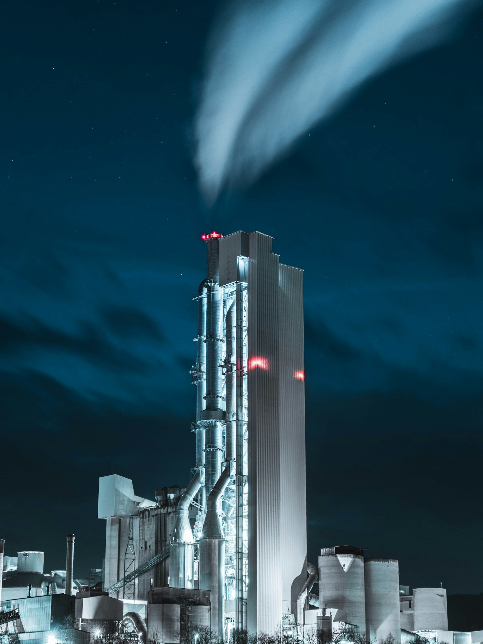 Night view of a large industrial factory with tall chimneys releasing smoke or steam, illuminated with bright lights at the base, against a dark sky with visible stars.
