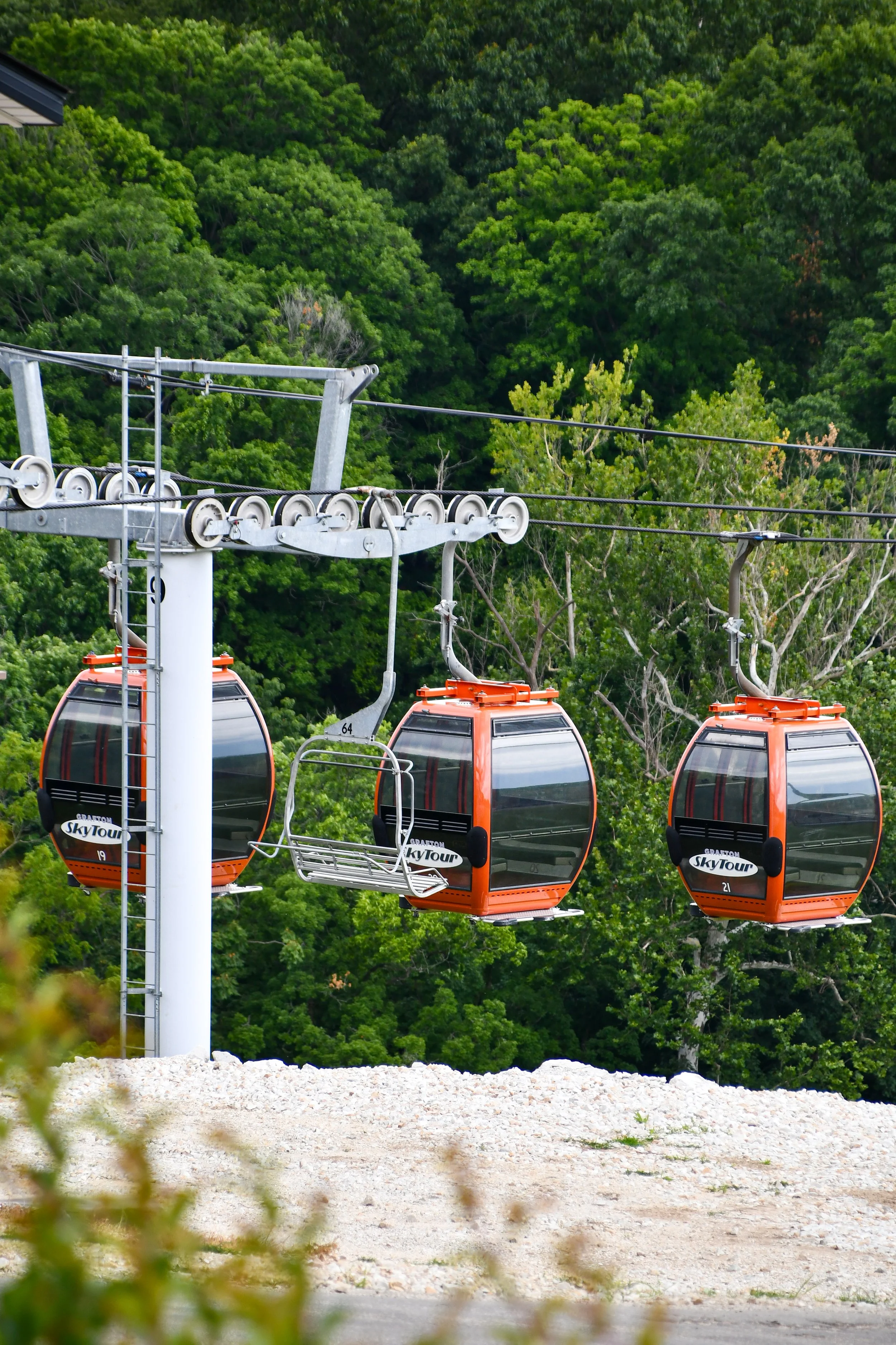 Multiple orange gondola-style cable cars hanging from a cable over green trees, with a white support pole in the foreground and a gravel area below. Aerie's in Grafton, Illinois.