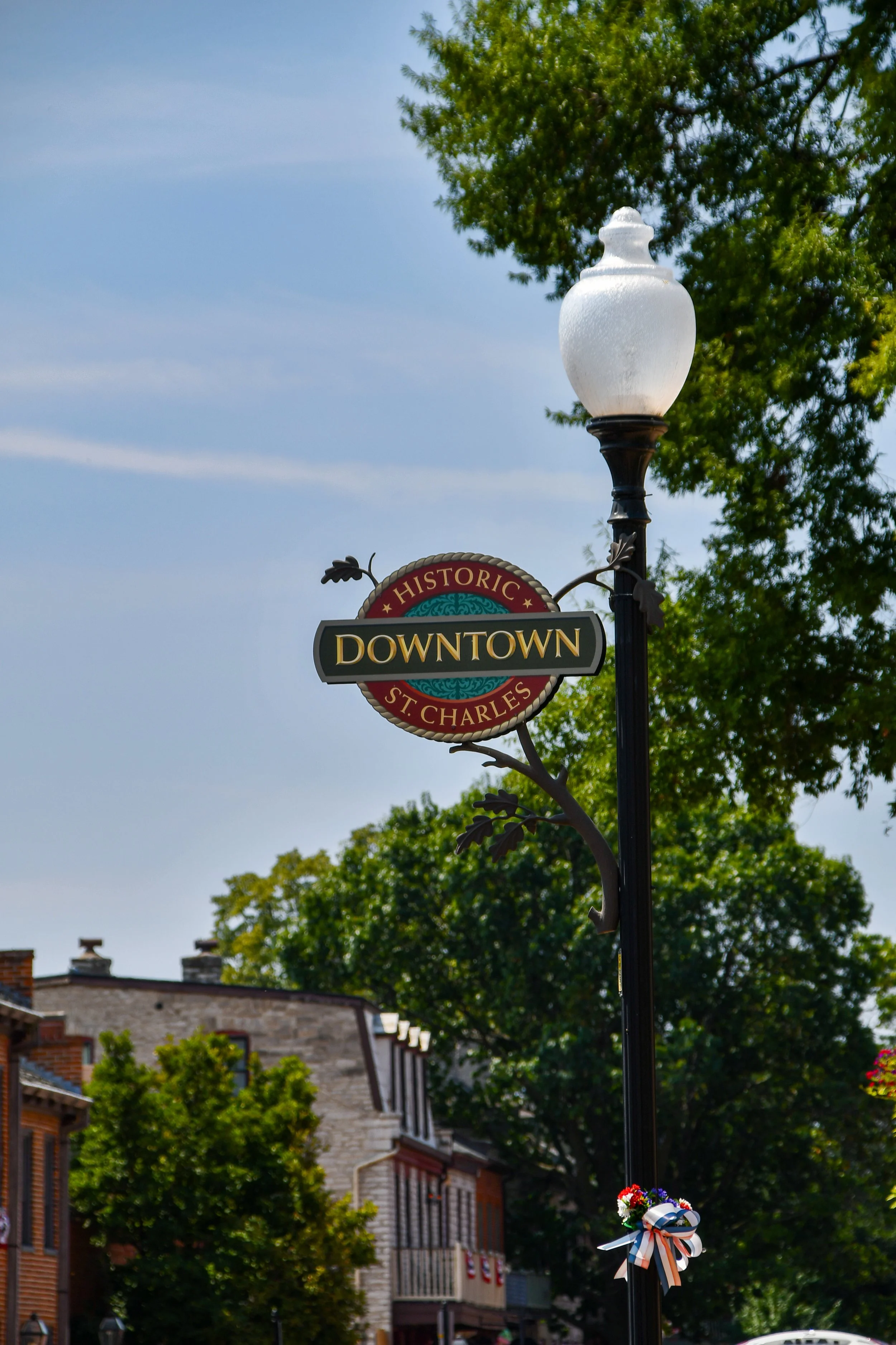 A streetlamp with a white glass globe on a black pole, decorated with a ribbon and flowers, and a sign reading 'Downtown' and 'St. Charles,' in an urban area with trees and brick buildings in the background.