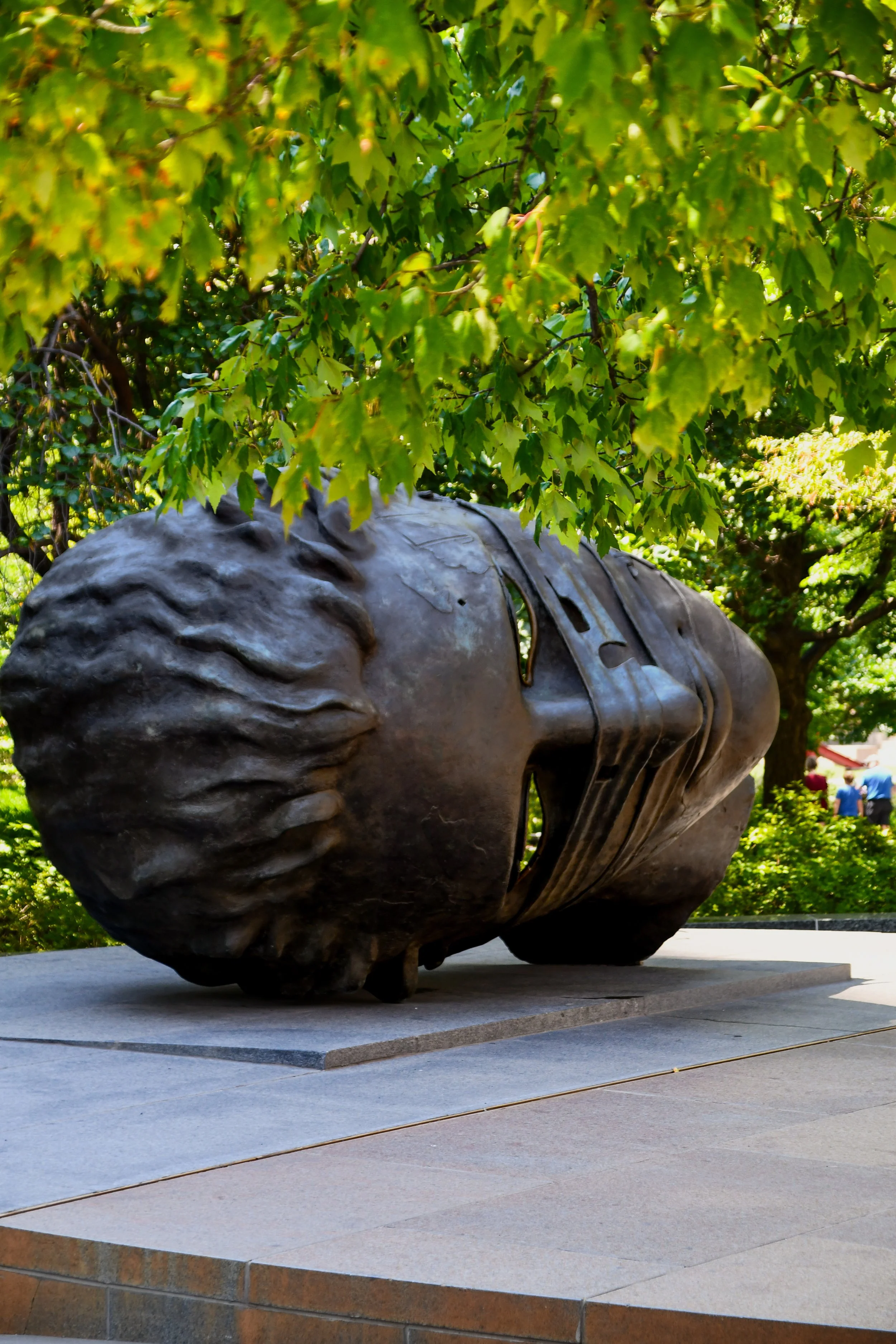 Large bronze sculpture of a human head lying on its side, with a face and detailed hair, set outdoors on a stone platform surrounded by lush green trees. Citygarden in St. Louis.