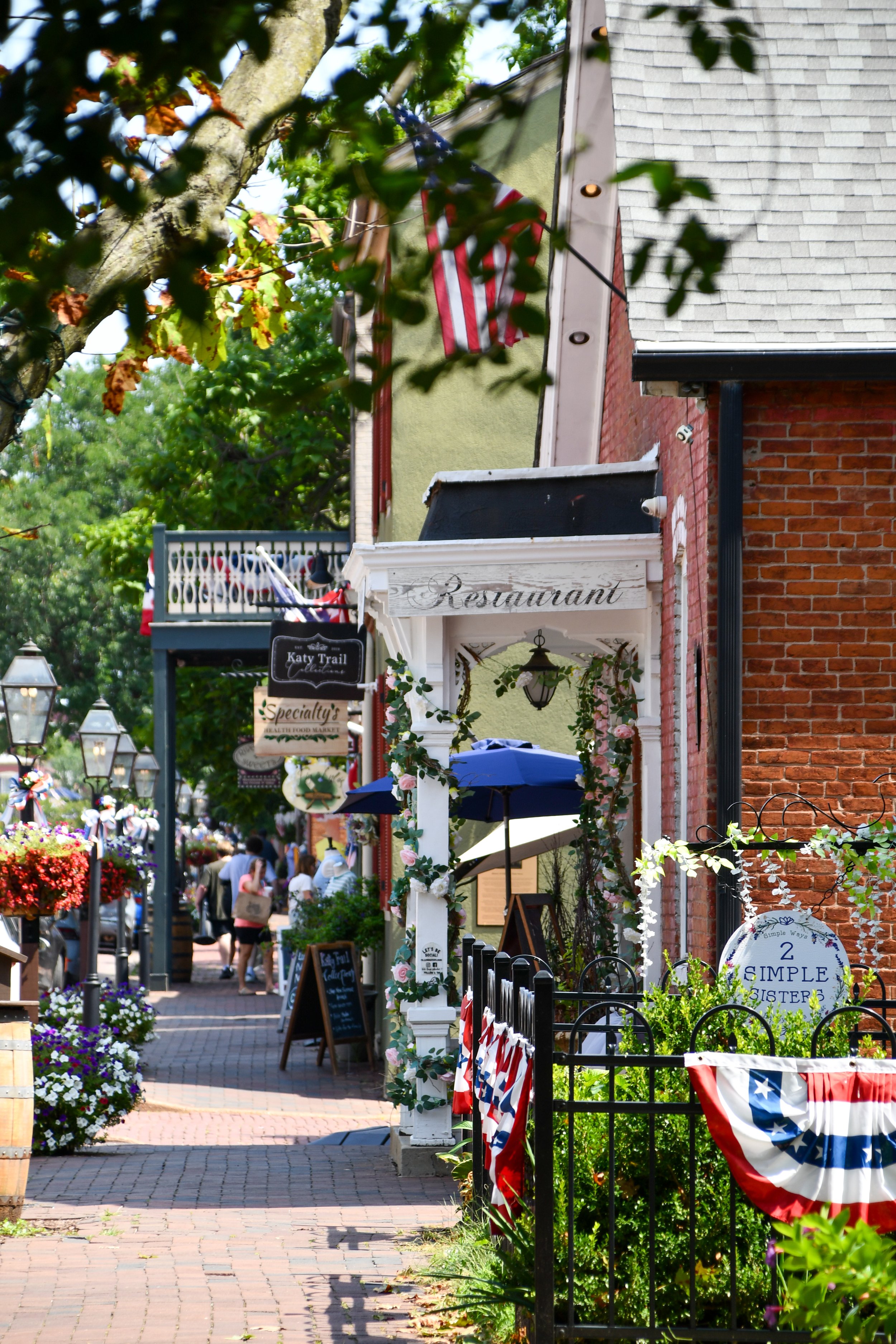 Sidewalk shopping street decorated with American flags, patriotic bunting, and flowers. People are walking and storefronts include a restaurant and specialty store, with flowers and hanging signs. Downtown Historic St. Charles District.