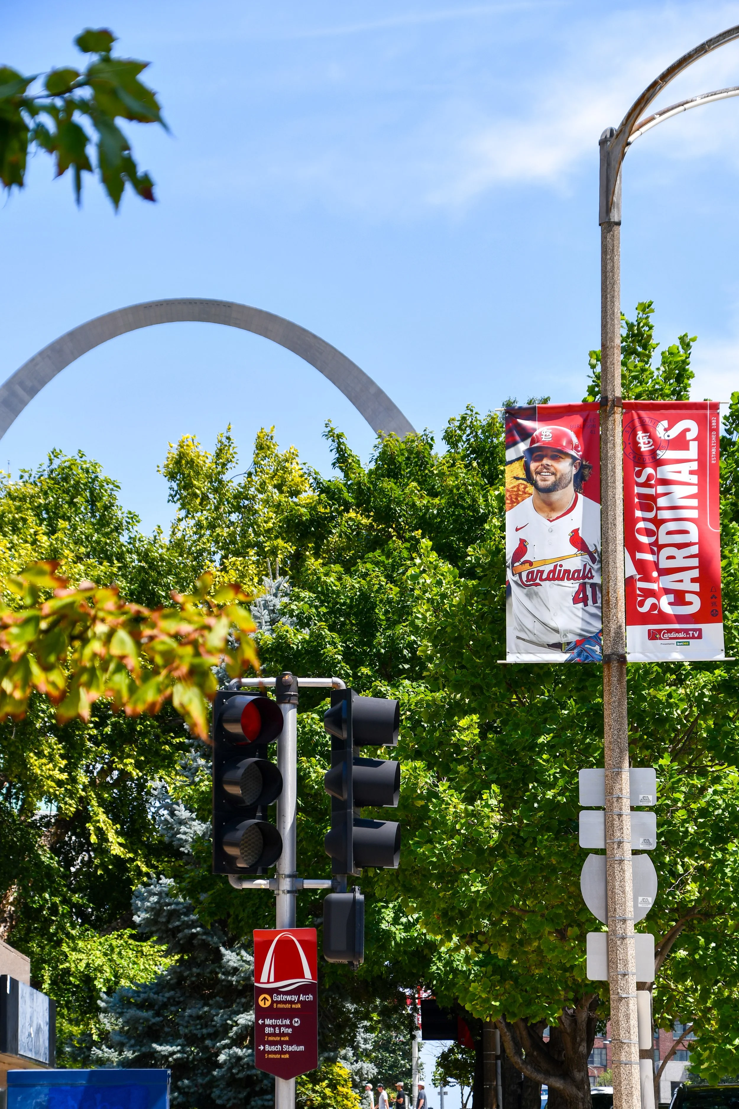 A street view featuring green trees, a traffic light, a banner with a baseball player in a cardinal jersey, and a sign pointing towards Gateway Arch, with a partly cloudy sky in the background. Downtown St. Louis and Cardinals banner.