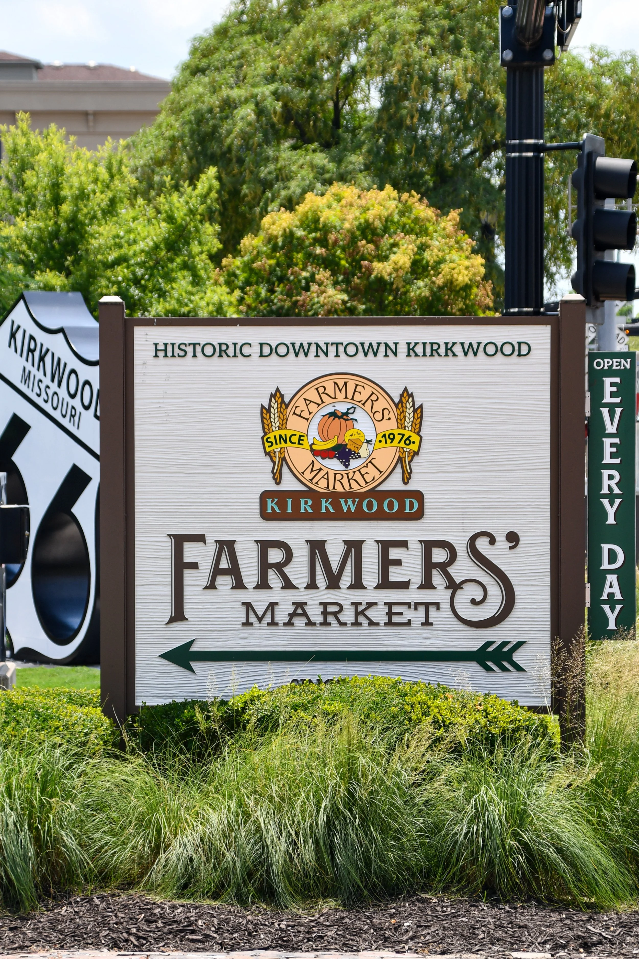 Sign for Farmers' Market in Kirkwood, Missouri, indicating it's open. There are green trees and bushes in the background, along with a traffic light.