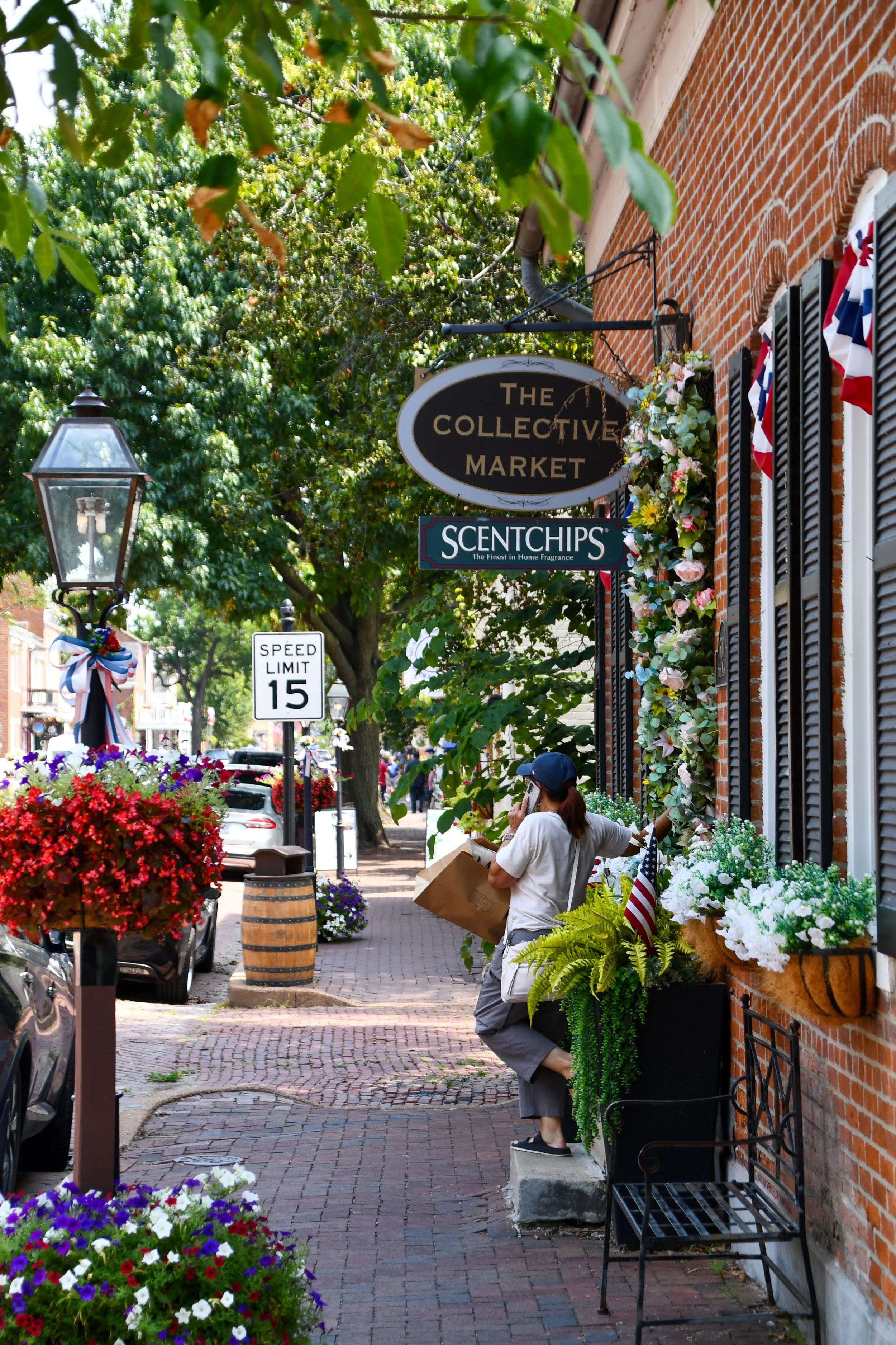 A woman standing on a sidewalk in front of a brick building with flower boxes, picking flowers. The scene is decorated with patriotic ribbons and flags, with a sign reading 'The Collective Market' and another advertising scented candles. There are cars parked along the street and a streetlamp. The weather is sunny and the street appears lively.