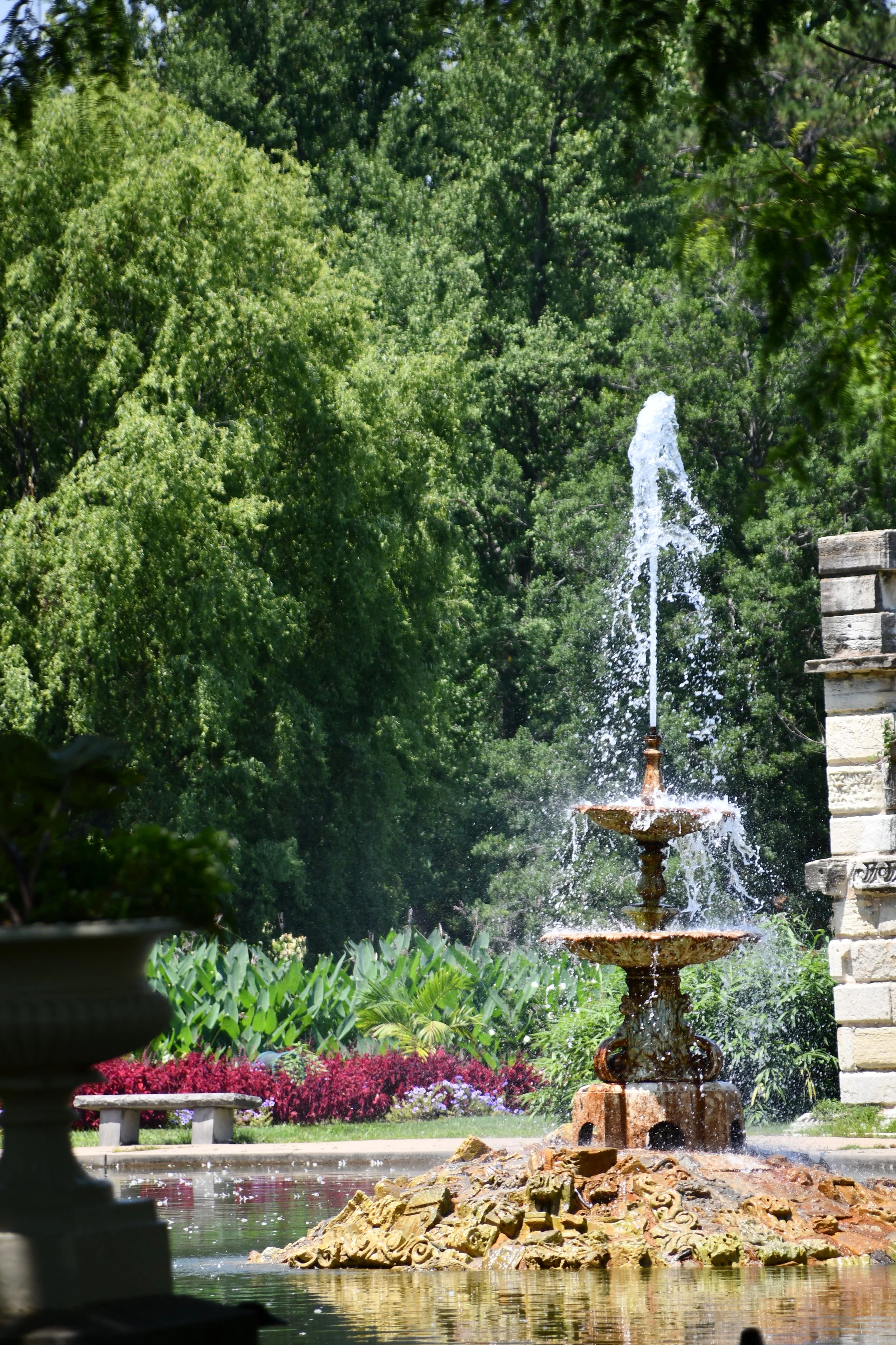 A decorative fountain in a garden with lush green trees, colorful flowers, and rocks around the fountain's base. Tower Grove Park in St. Louis, MO.