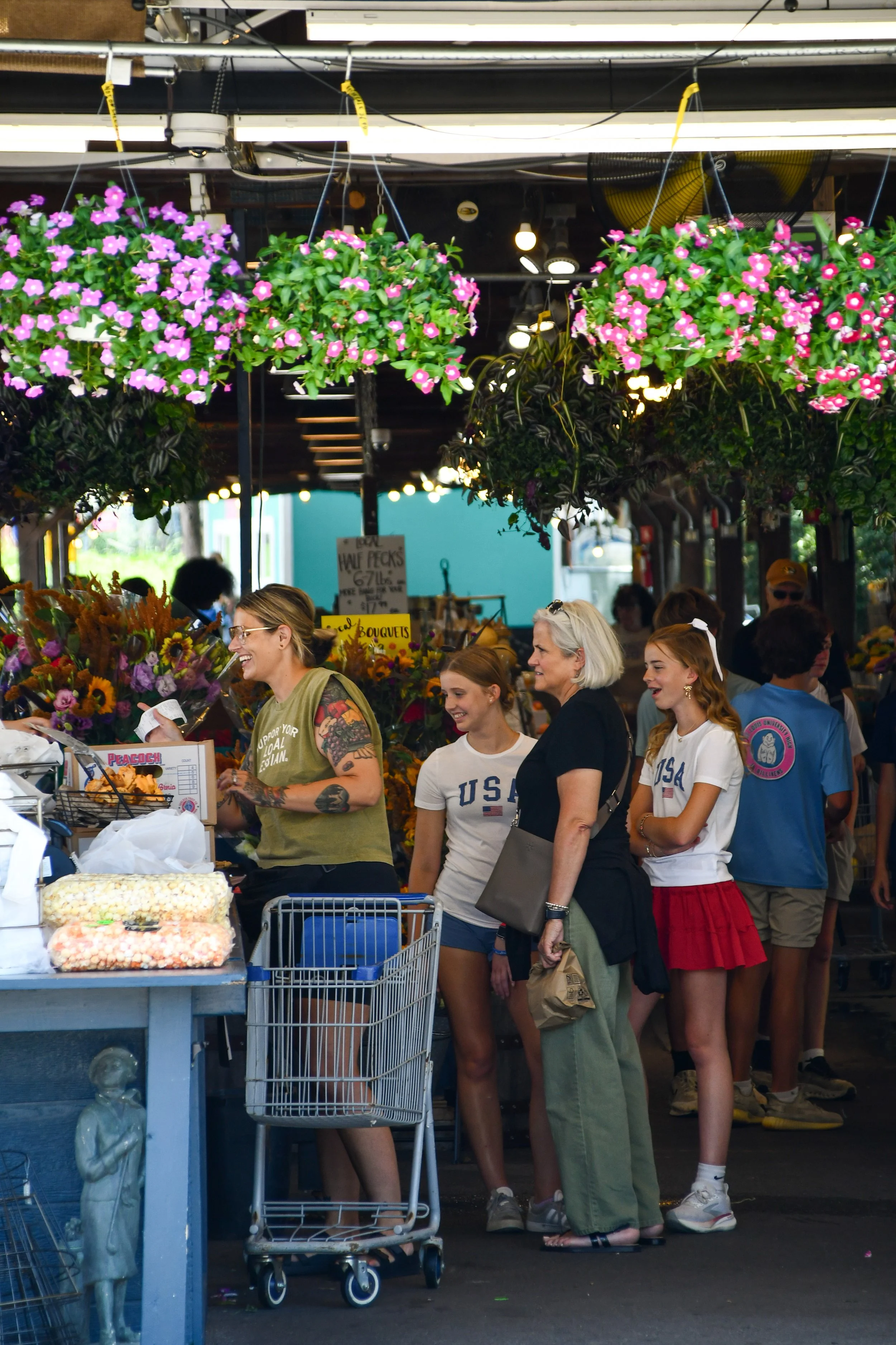 People shopping at an outdoor market with hanging flower baskets and various vendors selling items.