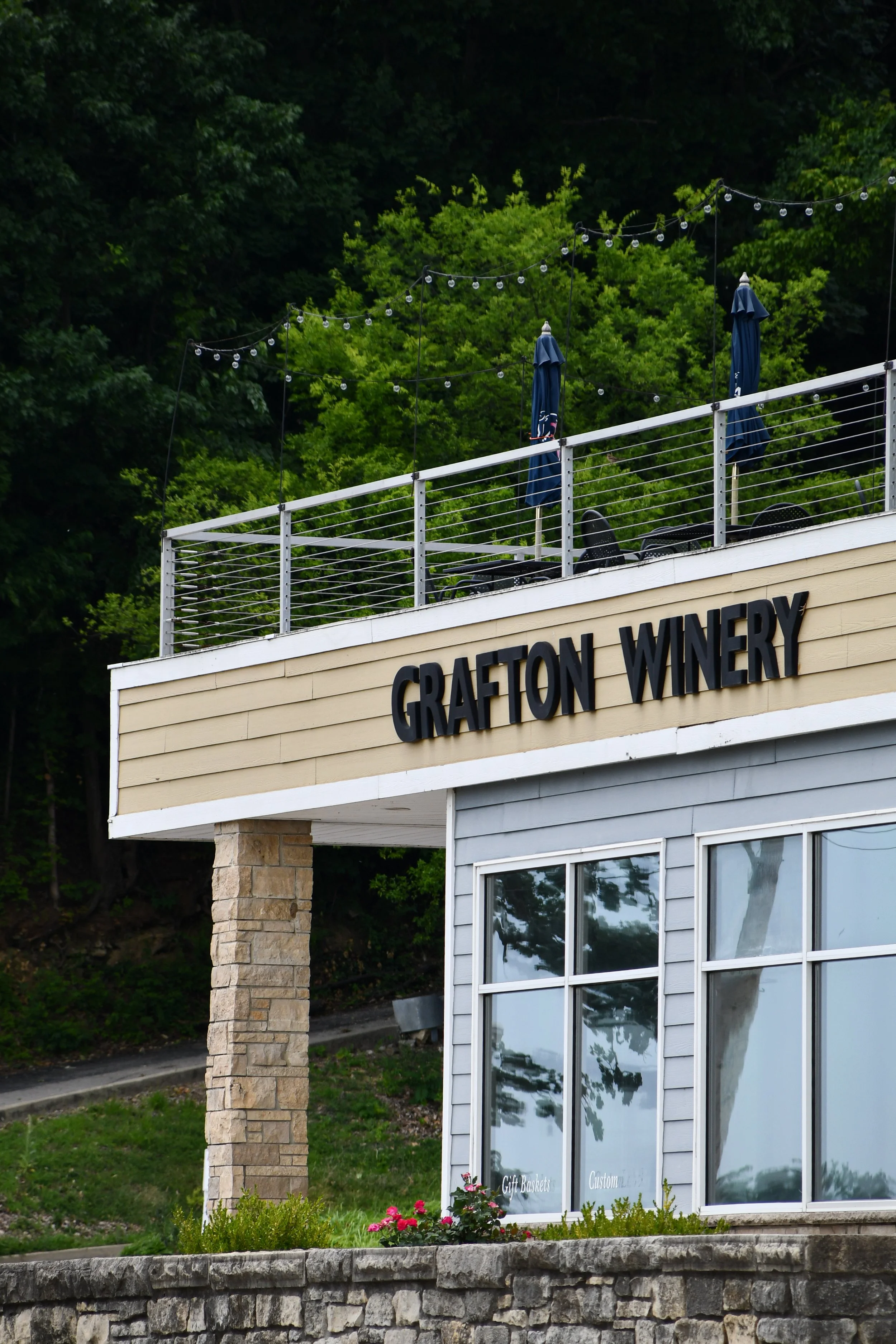 Exterior of Grafton Winery with a balcony having string lights, two blue patio umbrellas, and a glass-fronted shop window with flower pots outside, against a backdrop of green trees.